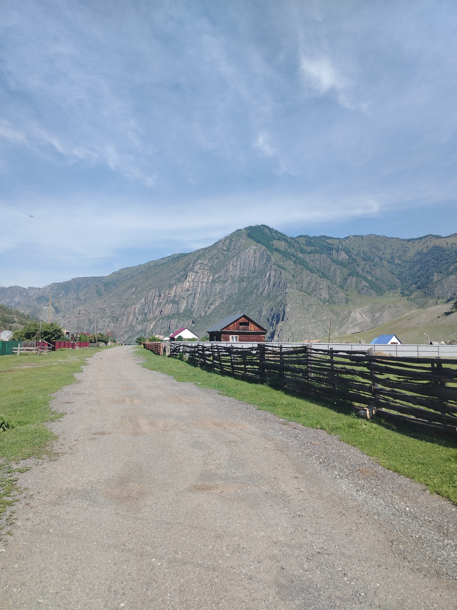 The image depicts a rural scene in Iodro, Russia, on a clear day with a few wispy clouds in the blue sky. A dirt road stretches into the distance, flanked by green grass and a rustic wooden fence. On the right side of the road, a cluster of houses can be seen, including a prominent wooden house with a red roof and a smaller building with a blue roof further down. Beyond the village, a majestic, rugged mountain range dominates the background, its slopes covered in sparse vegetation and rock faces. The overall atmosphere is serene and tranquil, suggesting a remote, mountainous region.
