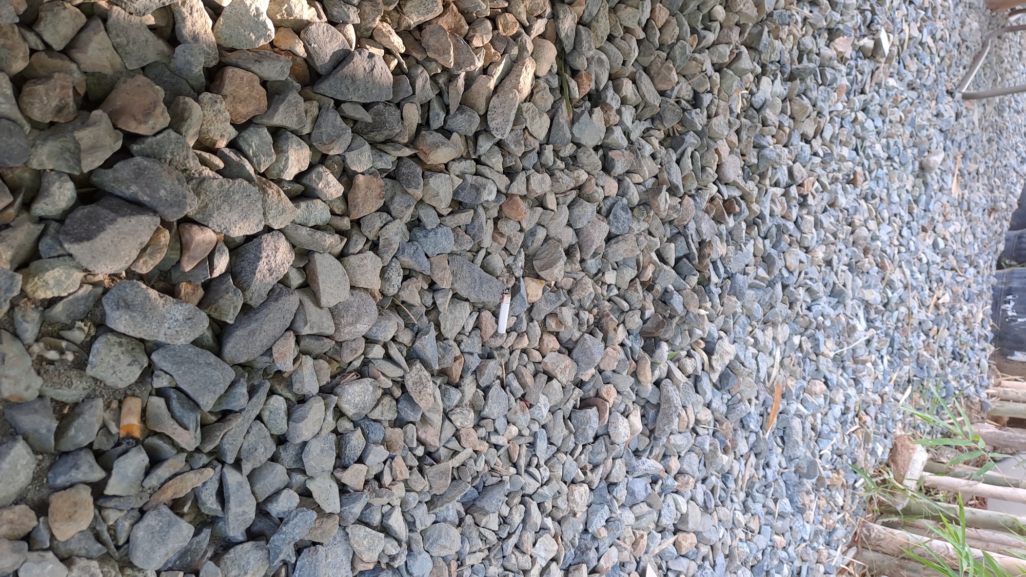 The image shows a close-up, ground-level view of a gravel path in Bireuenmeunasahdayah, Indonesia. The gravel consists of a mixture of gray, tan, and brown stones of varying sizes, some sharp and angular, others more rounded. Sunlight casts shadows across the stones, creating a textured surface. In the mid-ground, a white cigarette butt with an orange filter is visible, lying on top of the gravel. In the background, the blurry shapes of black planters or large pots can be seen, along with some thin, vertical stems and leaves, suggesting vegetation. A metallic, leg-like structure, possibly part of a chair or table, is also visible in the upper left corner. The overall impression is one of an outdoor area, possibly a garden or pathway, with some litter present.