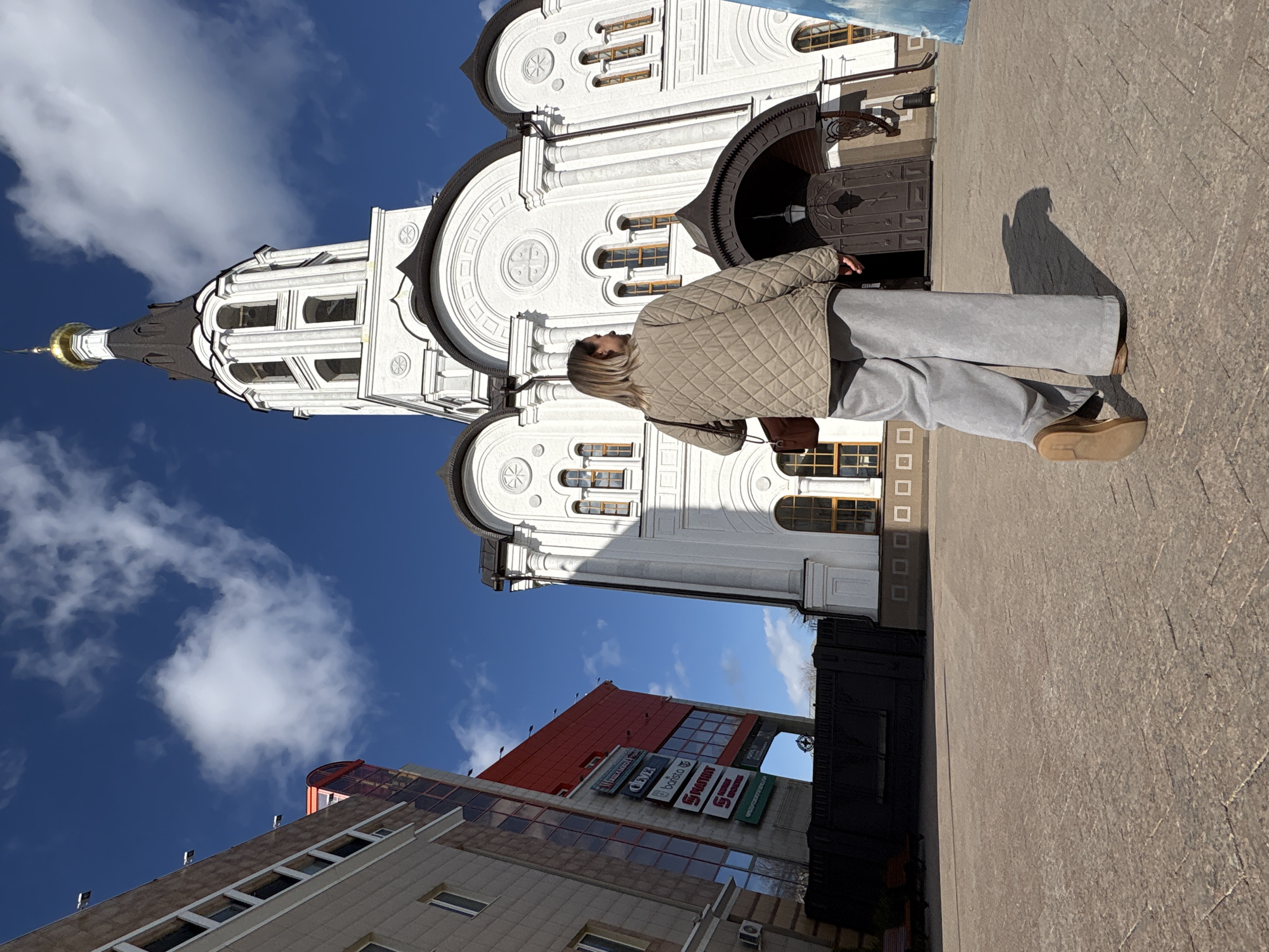 A woman in a beige quilted jacket and gray pants stands facing a white church with a black dome and gold spire against a blue sky with scattered clouds. The church has rounded arches and decorative circular designs. The woman is positioned to the side of the church entrance, her back mostly to the viewer. Below the church, portions of other buildings are visible, including a building with multiple store signs in Russian. The ground is paved. The photo appears to have been taken at an angle. Based on the architecture and store signs, the location is Ulyanovsk, Russia.
