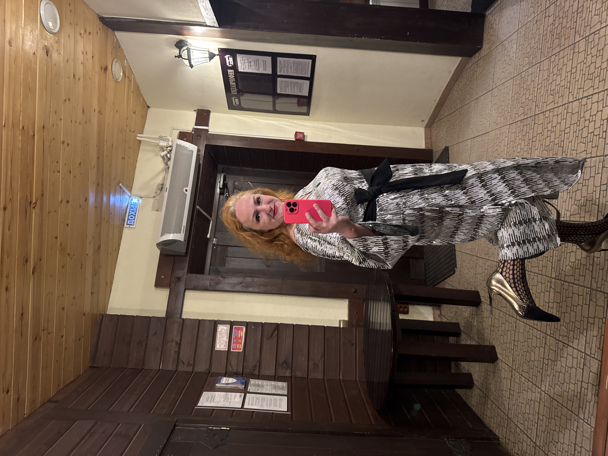 A red-haired woman is taking a selfie with a pink smartphone in the interior of a restaurant in Chaikovsky, Russia. She is wearing a patterned silver and black outfit with a black bow at the waist and black and gold high heels. The interior features wooden walls, a tiled floor, and a wooden table. A menu is displayed on the wall. An exit sign is visible.