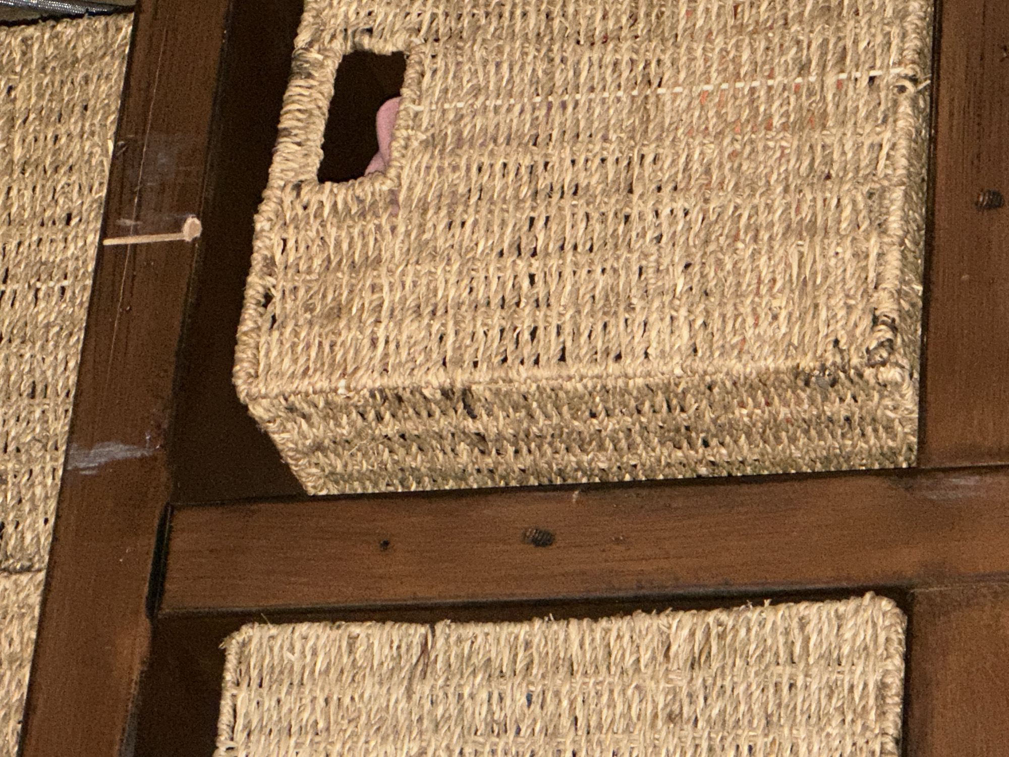 The image shows a close-up view of woven storage baskets on a wooden shelf. The baskets are made of a natural fiber, likely straw or seagrass, and have a textured appearance. One basket is partially visible on the left, and a larger basket with a rectangular opening is centered in the image. A piece of pink fabric is visible inside the centered basket. The wooden shelf appears to be dark brown. There are no people visible in the image. The scene is indoors, likely in a domestic setting. The location in the city of Thora, Australia, is not determinable from the visual information provided.