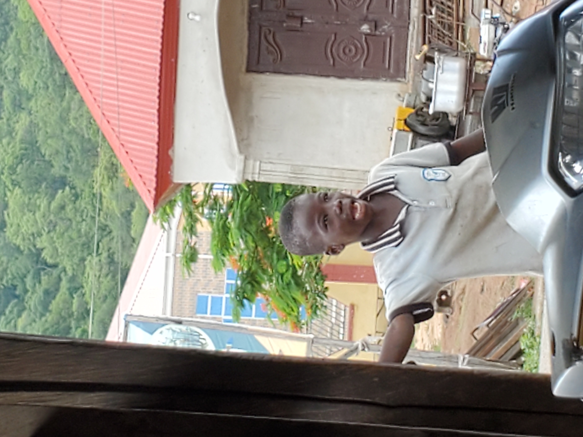 A boy wearing a light-colored, collared shirt with dark stripes on the collar and sleeves is standing next to a silver motorcycle in Mararaba, Nigeria. The background includes a building with a red roof and brown doors, trees, and another building with blue windows. The bottom of the frame is obscured by a dark wooden structure.