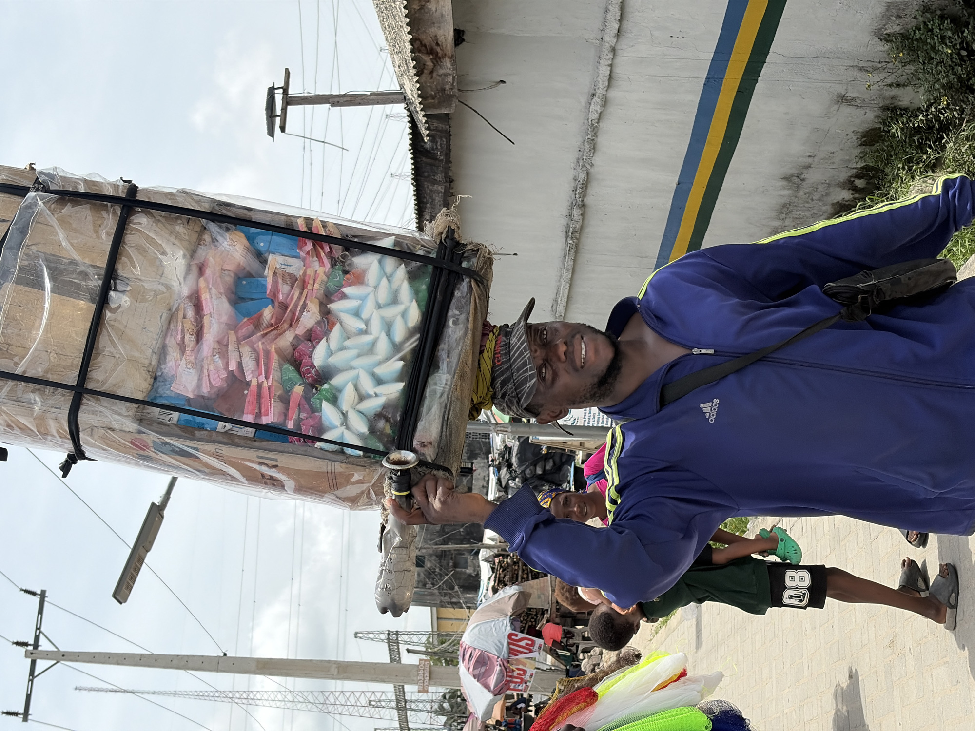 The image captures a man standing on a paved street in Igboefon, Nigeria, carrying a large, square package atop his head. The package is wrapped in clear plastic and contains a variety of items, including pink and blue paper goods and white, star-shaped objects. The man is wearing a blue Adidas tracksuit jacket, a patterned cap, and a black shoulder bag. He holds a small, dark object in his hand and looks towards the camera with a smile.
The background shows a bustling street scene with people and various goods for sale. A white concrete wall with blue and yellow stripes runs along one side of the street. Power lines and utility poles are visible overhead. The scene appears to be a busy market or commercial area in the city.