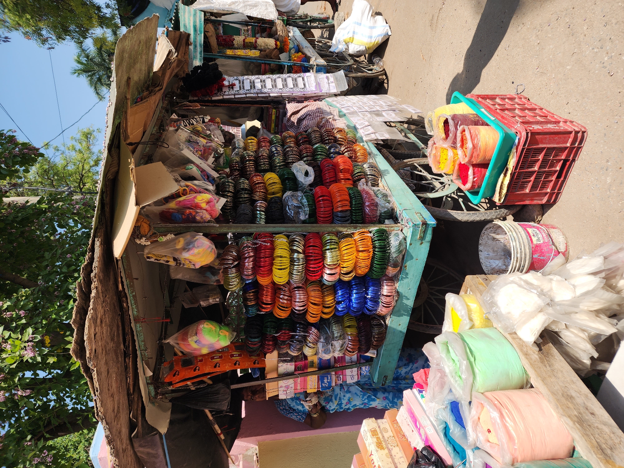 An outdoor market scene is depicted under bright daylight in the city of Unknown, Unknown. The primary subject is a rustic, light teal-painted wheeled vendor's stall with a weathered, makeshift roof constructed from various sheets of material, including corrugated metal and cardboard.
The stall is heavily laden with merchandise. The central display consists of numerous vertical stacks of multi-colored glass bangles, featuring shades of red, orange, yellow, green, blue, brown, black, and clear. Above and around the bangles, various items are suspended in clear plastic bags, including colorful plastic toys and other small wares. Flat orange, rectangular objects with embossed text are also visible hanging.
To the left, a woman wearing a blue and white patterned sari stands behind the stall, partially obscured by boxes and merchandise. She is actively engaged, holding an object in her hands. On a wooden surface to her left, several cardboard boxes, some pink and some patterned, are stacked. Further left, several large, clear plastic bags filled with brightly colored powders (pink, light green, light blue, yellow) are displayed on a lower wooden shelf. Adjacent to these, smaller white packets are piled. A paint-splattered white bucket is on the ground.
To the right of the main stall, another vendor's cart is partially visible, displaying items such as garlands and coconuts. The lower right foreground shows a red plastic crate supporting a green plastic tray containing multiple smaller clear plastic bags filled with colored powders (red, orange, yellow, maroon).
The ground is a light-colored, uneven paved or compacted dirt surface. Strong sunlight is present, indicated by distinct shadows, including a human silhouette on the ground in the mid-right. The background features green trees with visible purple blossoms, a clear blue sky, and overhead utility lines. The overall environment suggests a bustling, open-air marketplace.