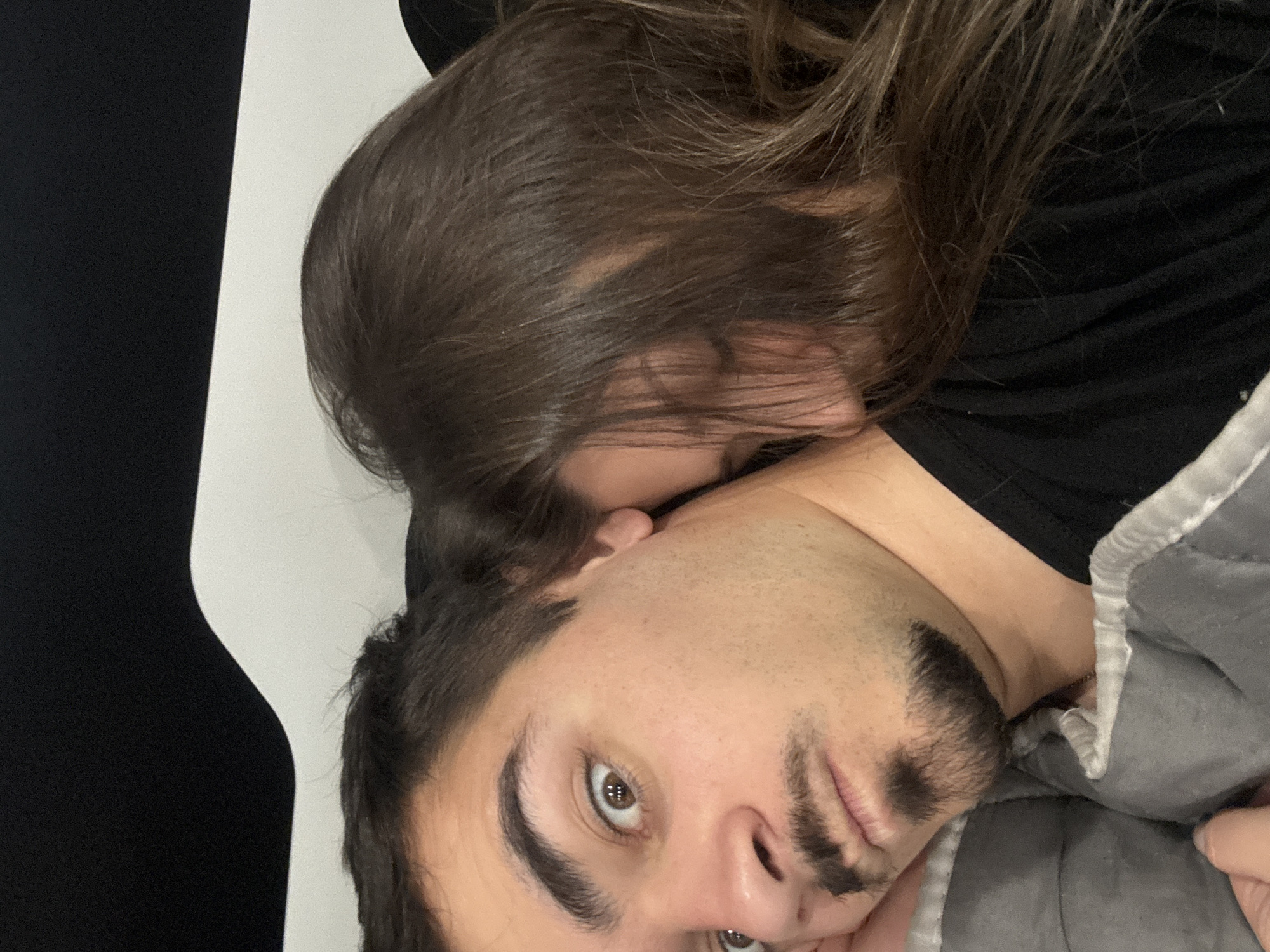 A man and a woman are lying together. The woman's head is on the man's neck, her face obscured by her hair. The man is looking directly at the camera. The background is a white wall next to a black wall. The location is unknown.