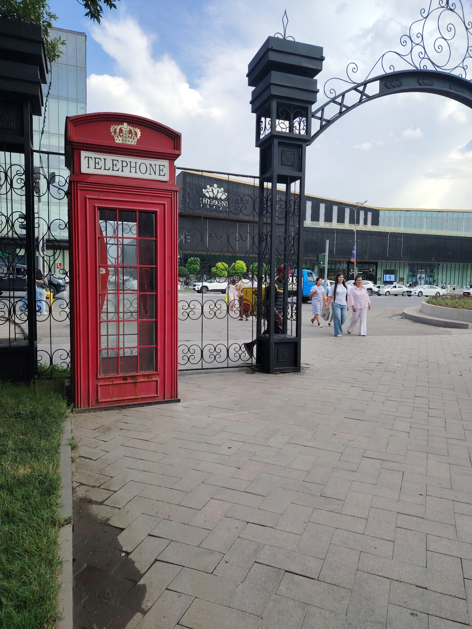 In Shymkent, Kazakhstan, a red British-style telephone booth stands on a paved walkway. Behind the booth, a black ornate metal gate with decorative scrollwork separates the walkway from a street where vehicles are present. Further in the background, a modern building with a glass facade and dark horizontal accents is visible. Several people are walking in the vicinity of the gate and on the street. The sky above is partly cloudy.