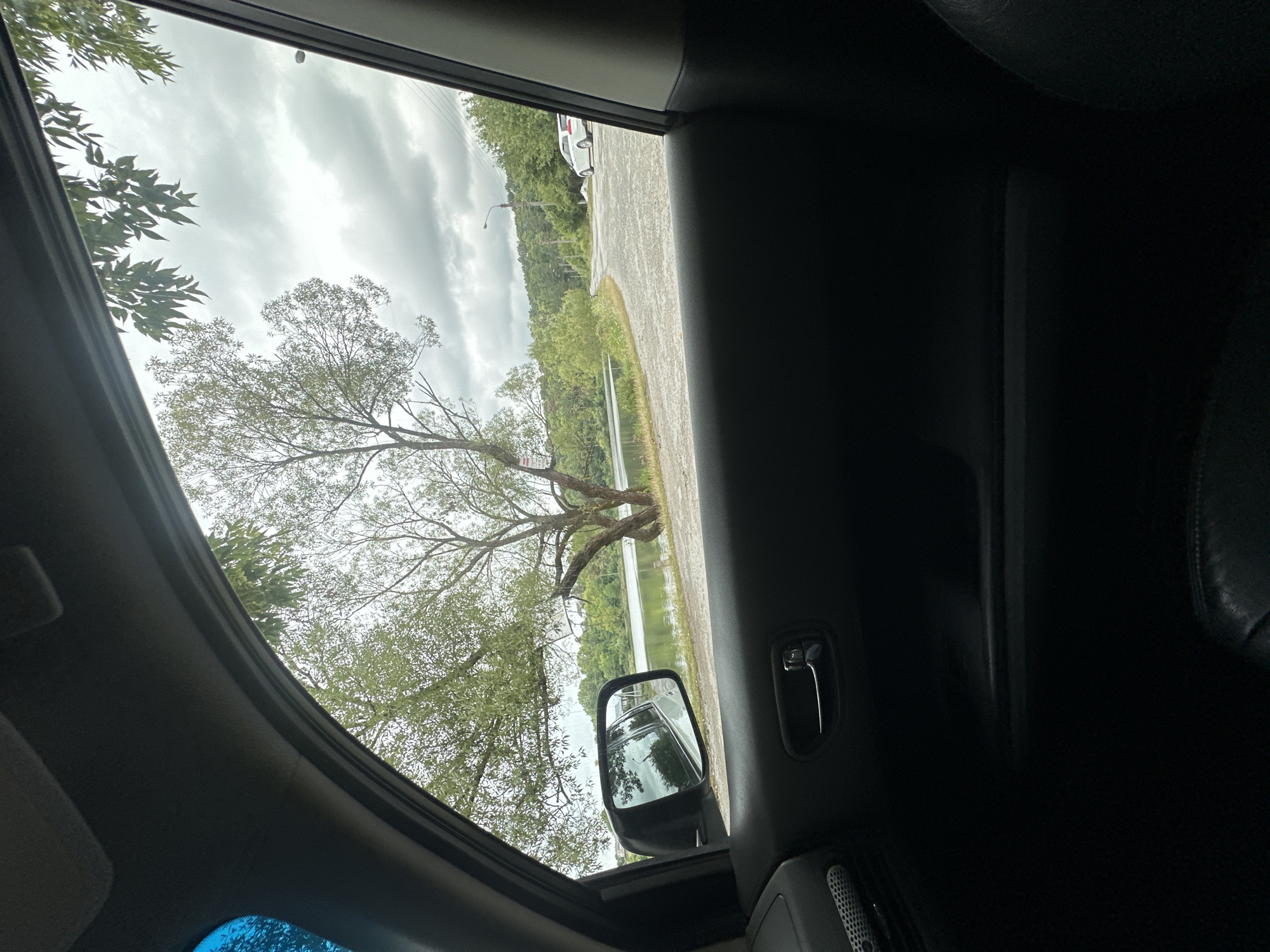 The image, taken from inside a vehicle, shows a view through the side window of a car. The foreground is dominated by the car's interior, including the door panel, door handle, and a portion of the seat. Outside the window, a body of water, likely a lake or river, is visible, bordered by lush green vegetation and trees. A large tree with sparse foliage stands prominently near the water's edge. Beyond the water, a gravel road curves through a hilly landscape covered in trees. A white car is parked on the side of the road in the distance. The sky is overcast with heavy clouds, suggesting a cloudy or rainy day. The scene appears to be set in a rural or semi-rural location, possibly on the outskirts of Makiv, Ukraine, given the geographical context.