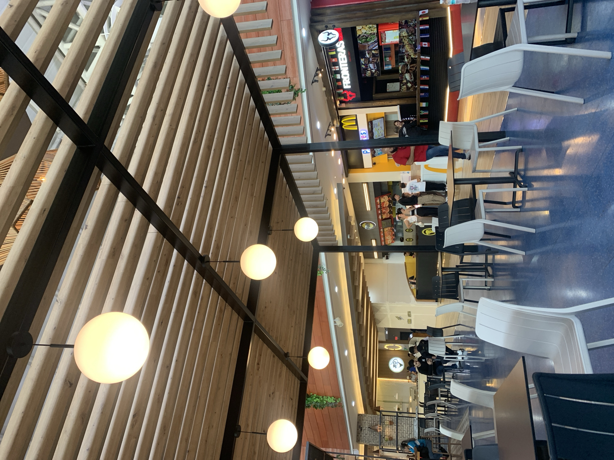 This indoor shot shows the food court inside the Sambil Margarita Mall in Porlamar, Venezuela. Viewed from above, the space is filled with fast-food restaurant fronts, seating areas with white plastic chairs, tables, and people eating. Above, there's a ceiling structure of wooden slats, with spherical lights hanging.