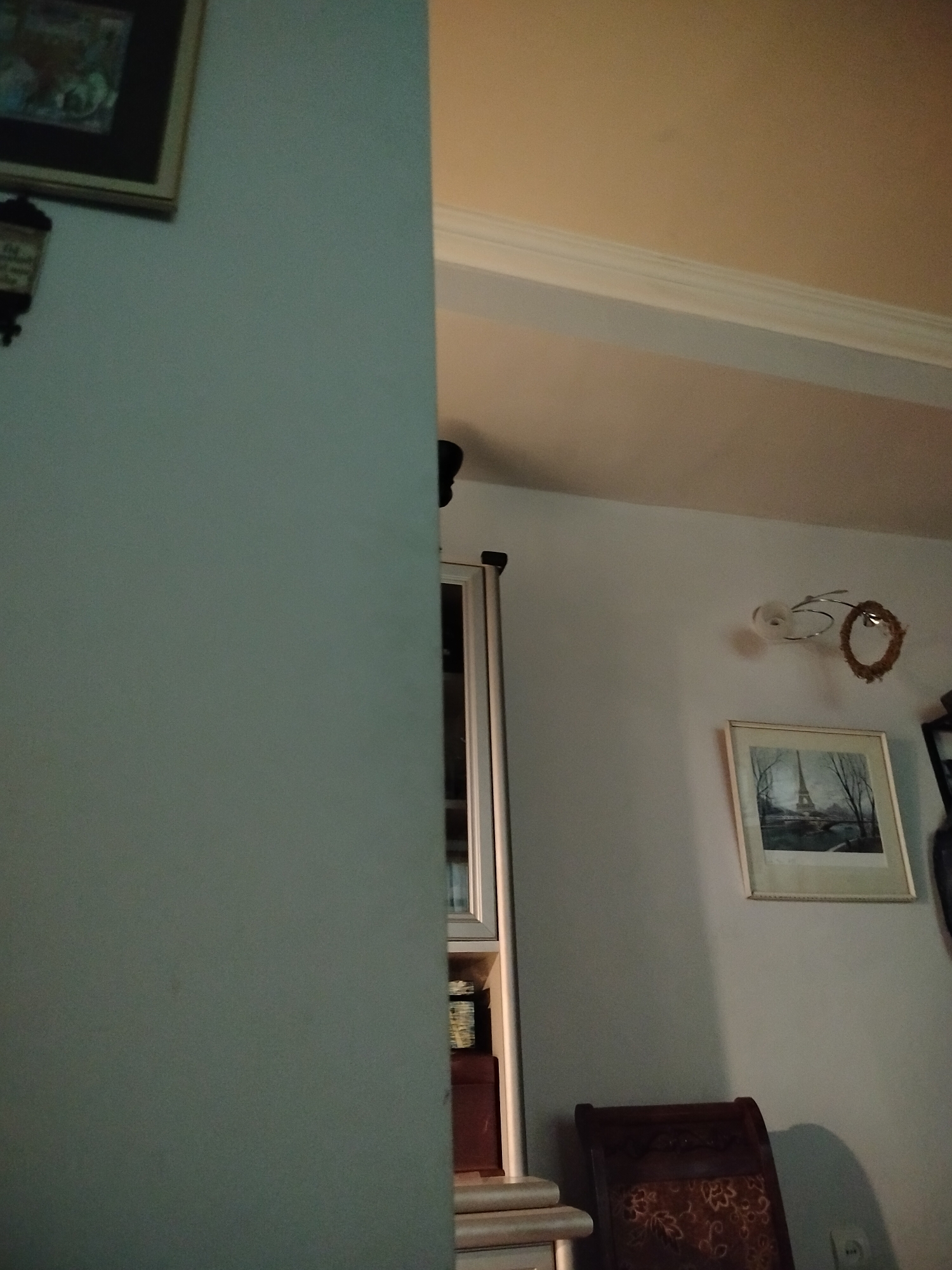 The image depicts an interior setting, likely a home, possibly located in Yerevan, Armenia. Dominating the left side of the frame is a partially visible wall painted in a muted color. A framed picture and a wall-mounted lamp are affixed to the wall.
The right side of the image reveals a dining area. A wall-mounted light fixture, a framed picture of the Eiffel Tower, and a wooden chair with patterned upholstery are noticeable. A partial view of a cabinet can be seen as well.
The environment appears dimly lit, suggesting an indoor setting with limited natural light.