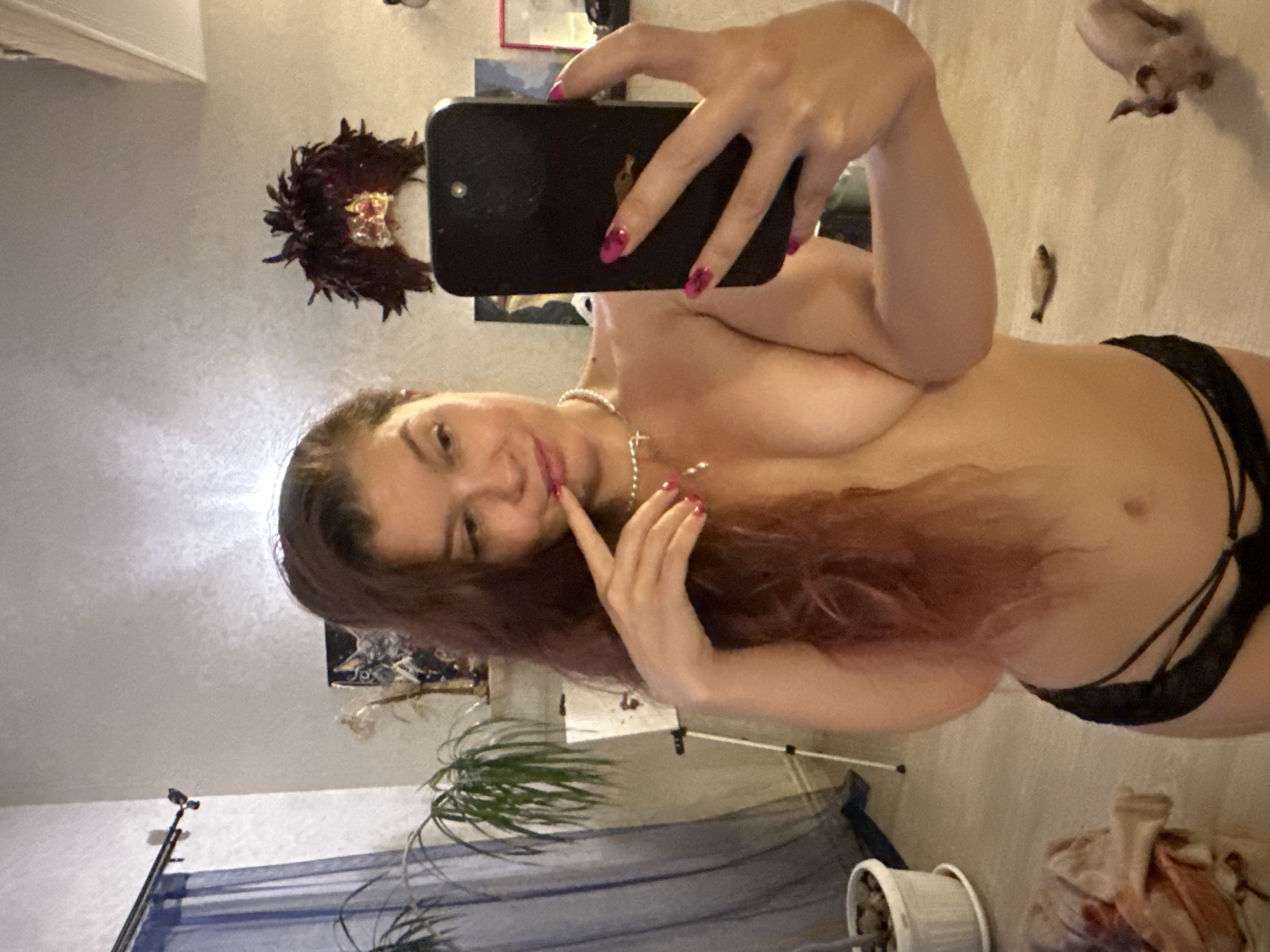 A woman is taking a mirror selfie in what appears to be a home environment, possibly an apartment, with eclectic decor. She is partially undressed, wearing only black lingerie bottoms. Her hair is long and dark, falling over her shoulder. She is holding a black smartphone in her right hand to take the photo. She has bright pink nail polish on her fingernails. She is wearing a pearl necklace. In the background, there are curtains, a potted plant, an easel with a painting, and what looks like a small animal in the foreground on the right, possibly a cat or a small dog. The overall lighting suggests it is indoors, with some artificial light present. There is no visual information within the image that allows for the identification of the city of Vladimir, Russia.