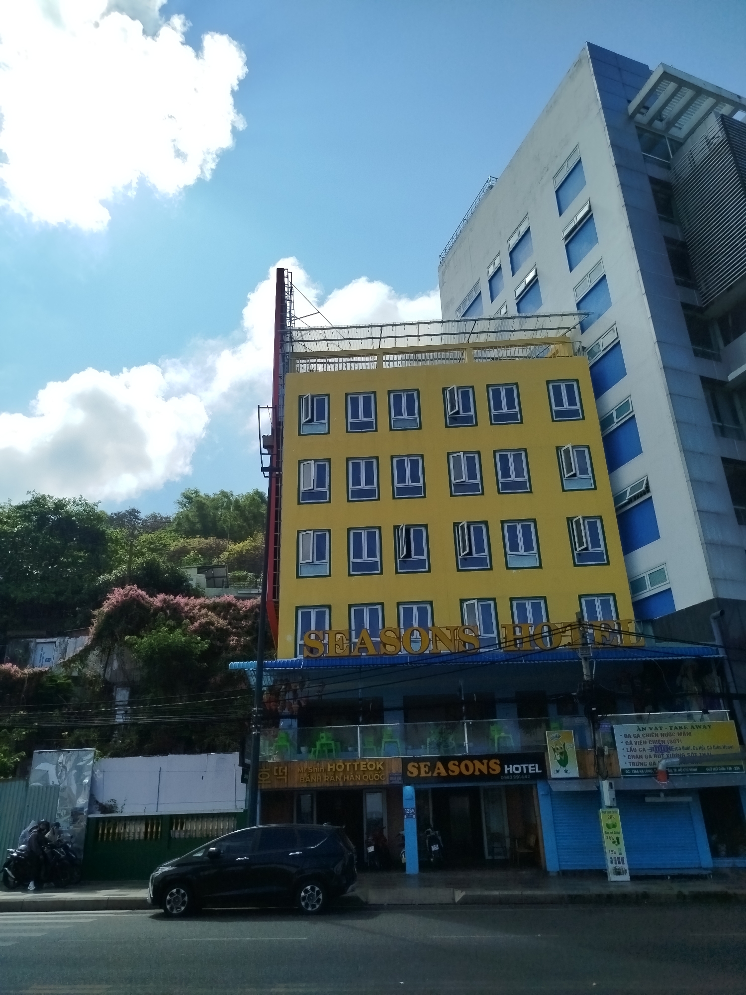 The image captures the Seasons Hotel located in Ho Chi Minh City, Vietnam. The hotel features a bright yellow facade with blue-trimmed windows, topped with a rooftop structure. Next to the hotel is a taller, white building with blue accents. The foreground shows a dark car parked on the street, with scooters parked nearby. In front of the hotel, there are various food stalls and signs, including a sign for "Mishin HOTTEOK," advertising Korean pancakes. Other signs advertise local food options. The surrounding environment includes a tree-covered hill in the background and a partly cloudy sky.