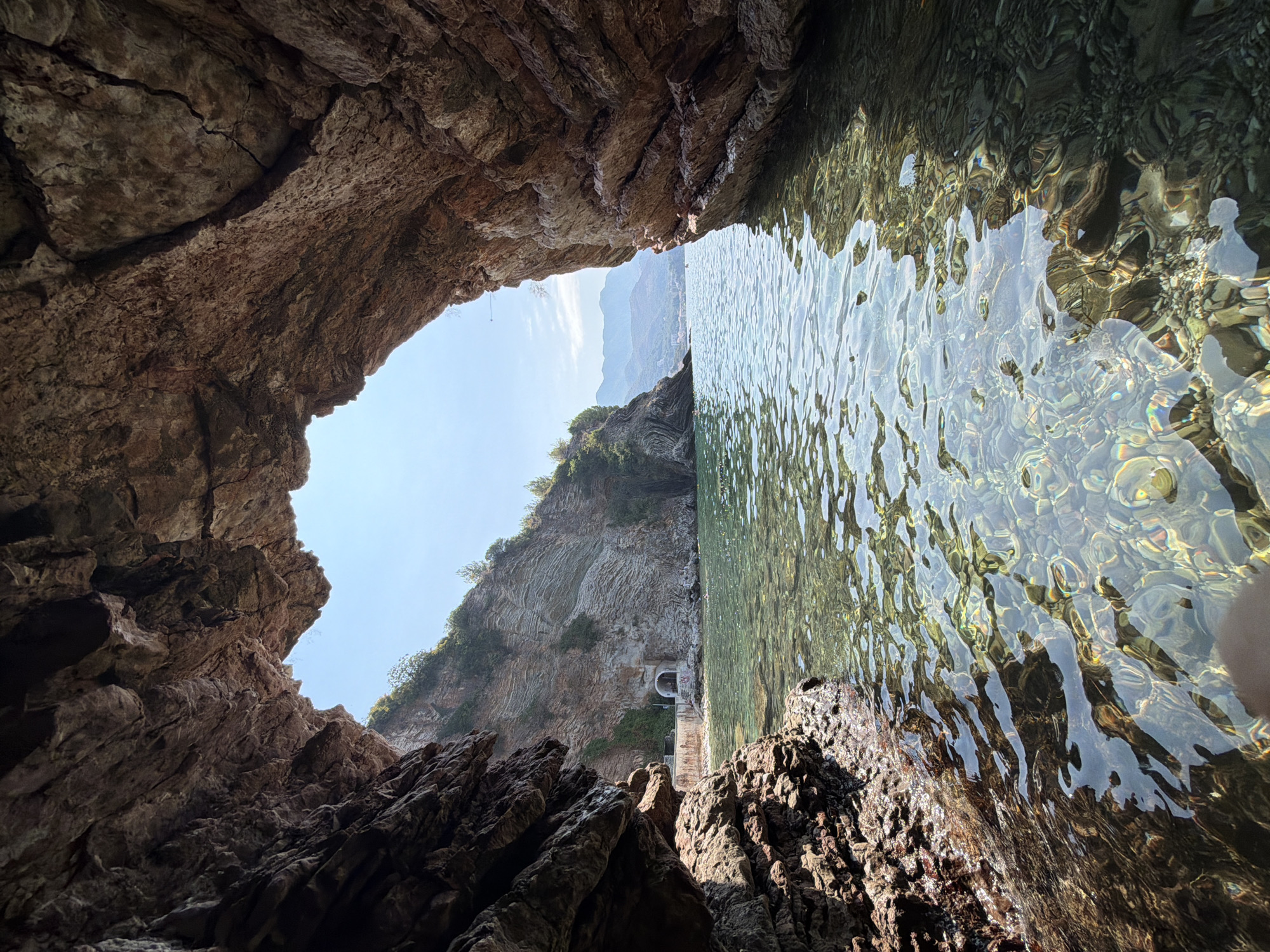 The image captures a view from within a sea cave, looking out towards a coastal landscape under a clear blue sky. The foreground is dominated by the rough, textured rock formations of the cave entrance, with sunlight illuminating the water. The water itself is remarkably clear, revealing a rocky seabed and creating dappled light patterns on its surface.
Beyond the cave's opening, a small, pebble beach is visible, backed by a steep, layered rock face. Embedded in this rock face is a light-colored archway or tunnel entrance, suggesting human activity or a man-made structure. Small structures with arched openings are visible near the beach.
Further in the distance, the coastline curves, with more rocky cliffs covered in sparse green vegetation. Mountains form the backdrop, their slopes softened by distance and atmospheric haze. The sea extends to the horizon, where it meets the sky.
The overall impression is one of a serene natural environment with a touch of human infrastructure. The scene appears to be located in the vicinity of Boreti, Montenegro, a coastal area known for its natural beauty and seaside attractions. There are no discernible people in the immediate foreground within the cave.