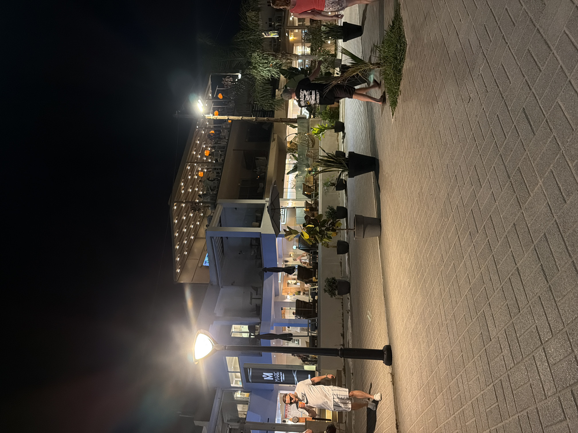 The image depicts a nighttime scene on a paved walkway in Georgioupoli, Greece, likely in a commercial or resort area. In the foreground, a man wearing a white t-shirt and shorts walks from left to right, holding a drink. A prominent streetlamp illuminates the walkway and the man. Behind him, a multi-story building with an open-air upper level is visible, featuring numerous lights and patrons dining or socializing. Several potted plants are strategically placed along the walkway. Further to the right, a couple walks away from the camera, with the woman in a pink top and patterned shorts, and the man in a black t-shirt and shorts. The overall atmosphere suggests a lively evening, possibly near a beach or a promenade, given the tropical-looking plants and open-air dining establishments.