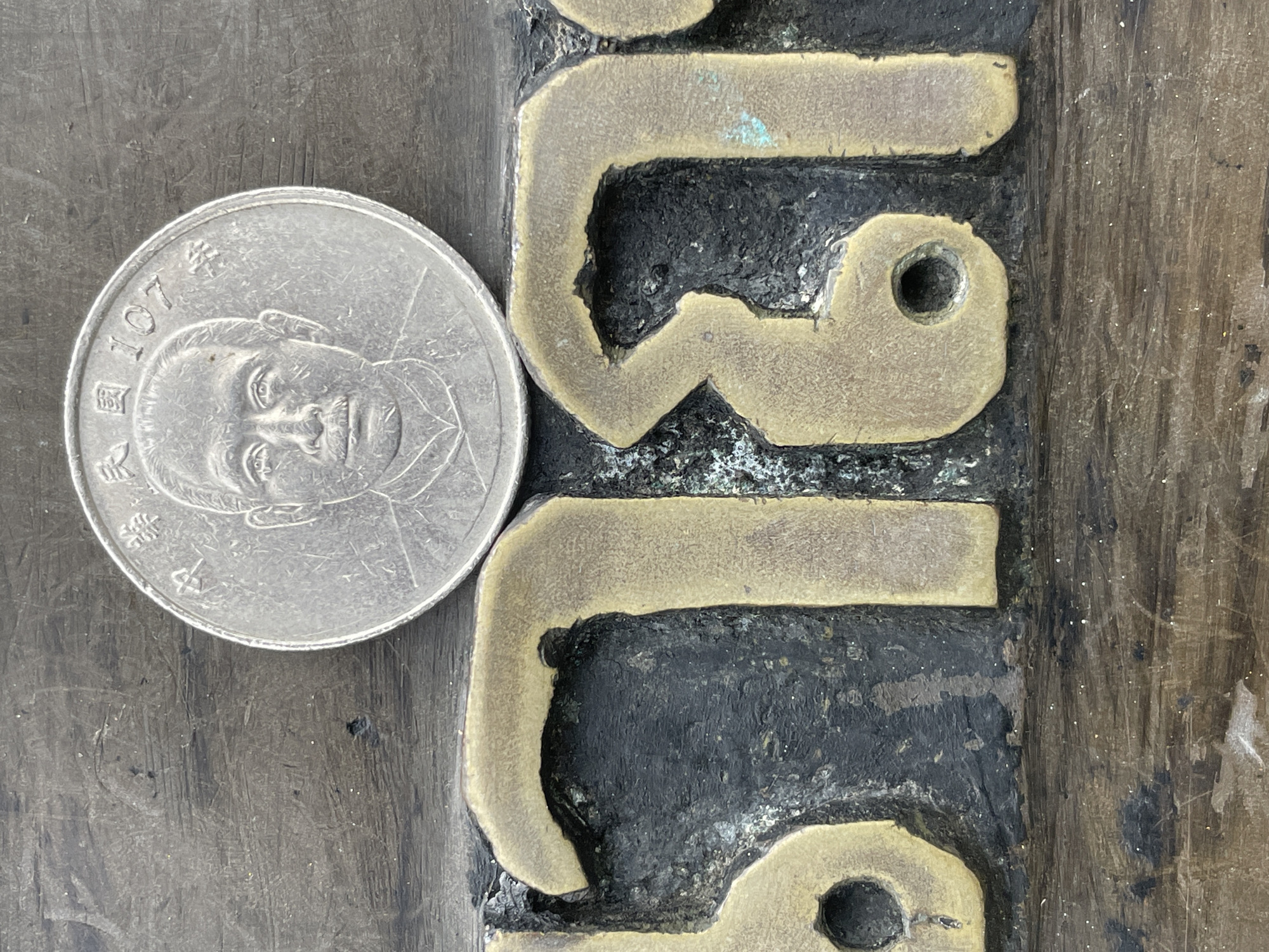 A close-up, top-down shot focuses on a silver coin resting on a dark, textured surface. The coin features a portrait of a man and Chinese characters, indicating it's a Taiwanese coin minted in the 107th year of the Republic of China. Below the coin, large, raised brass characters are embedded in the dark surface. The characters, which appear to be Thai script, are aged and show signs of oxidation, with some green patina. The overall environment suggests an outdoor setting, possibly on a plaque or monument. Based on the presence of Thai script and the context provided, this scene is located in Na Chom Thian, Thailand.