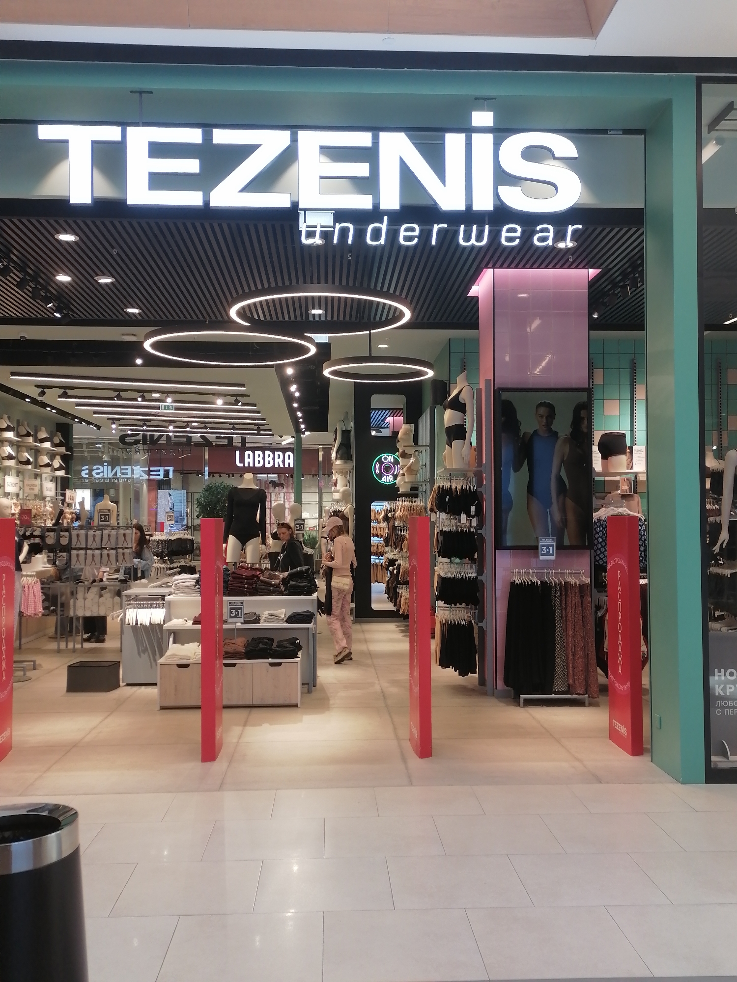 The image shows the storefront of a "TEZENIS" underwear store. The store is well-lit with bright white signage and internal lighting. The entrance is flanked by red pillars and displays of clothing. Inside, mannequins are dressed in underwear and clothing, and racks of garments are visible. Several people are present in the store, including shoppers browsing the merchandise and possibly store employees. The flooring is light-colored tile. The overall environment appears to be a modern retail space within a shopping mall. The signage on the right side of the store entrance, though partially obscured, appears to have Russian text, and the name "TEZENIS" itself is a European brand, indicating the potential location in Moscow, Russia.