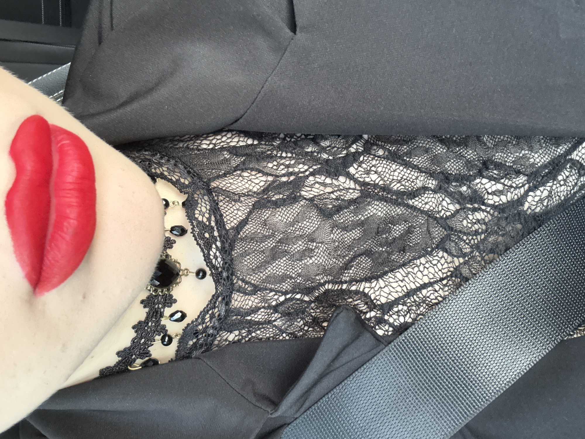 The image depicts a close-up view of a woman wearing a black lace top, a black blazer, and a necklace with black stones. The woman's skin is pale, and her lips are painted with bright red lipstick, pressed against her collarbone. A gray seatbelt crosses her body. The setting appears to be inside a vehicle. There is no information given to indicate the location is specifically Katunitsa, Bulgaria.