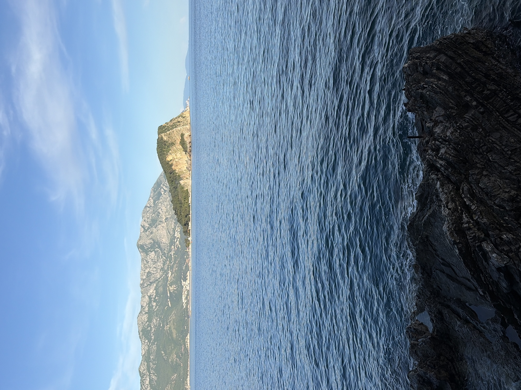 The image shows a view of the Adriatic Sea meeting the rocky coastline of Budva, Montenegro. The sky above is pale blue with some wispy clouds. A mountain range with vegetation, including some buildings on the summit, borders the sea on the left. The sea itself takes up most of the frame, and on the right, a dark, jagged rocky outcrop is visible. There are no people visible in the image.