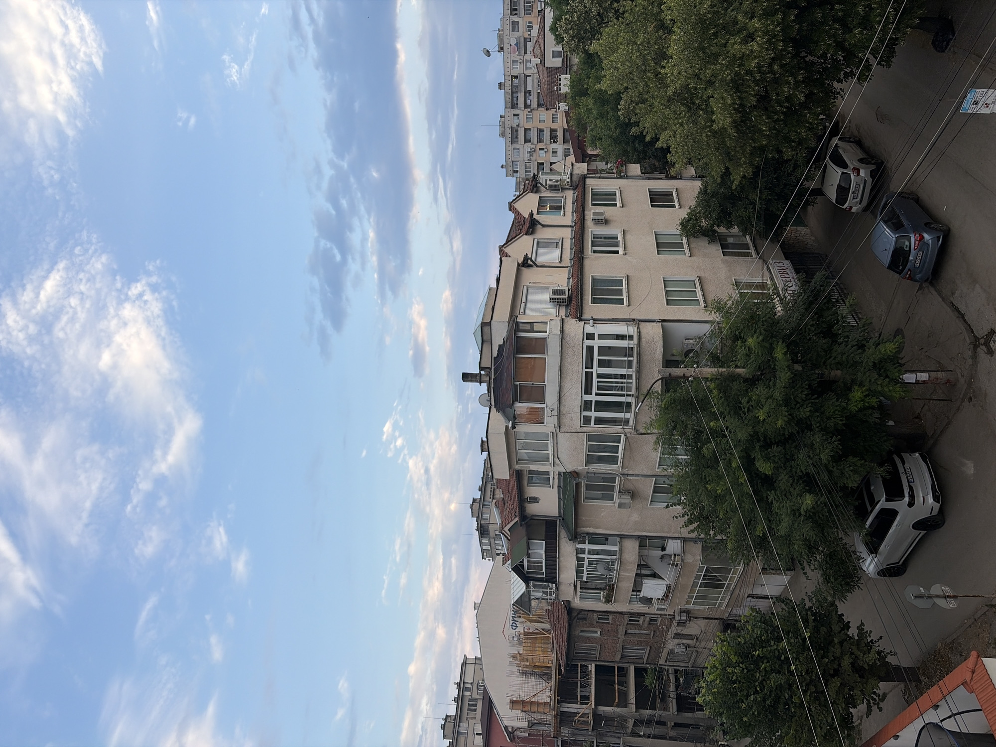 This is a vertically oriented image showing an urban scene. The left side of the image is dominated by a clear blue sky with wispy clouds, suggesting it might be daytime. The right side of the image features a row of multi-story apartment buildings, interspersed with large, green trees. A street runs alongside the buildings, and two cars are parked on it. One car is white and the other is dark blue or gray. Electrical wires are visible crisscrossing the scene. The architecture of the buildings appears to be from the mid-to-late 20th century, with various window styles and some with balconies or enclosed sunrooms. One building shows signs of ongoing construction with scaffolding and wooden structures visible. The overall impression is of a residential neighborhood, possibly in Eastern Europe, given the style of the buildings and cars. There is no clear indication of specific activity or emotion. The lighting suggests it could be late afternoon or early evening, with the sky still bright. There is some text on a sign visible on the street, but it is not legible.