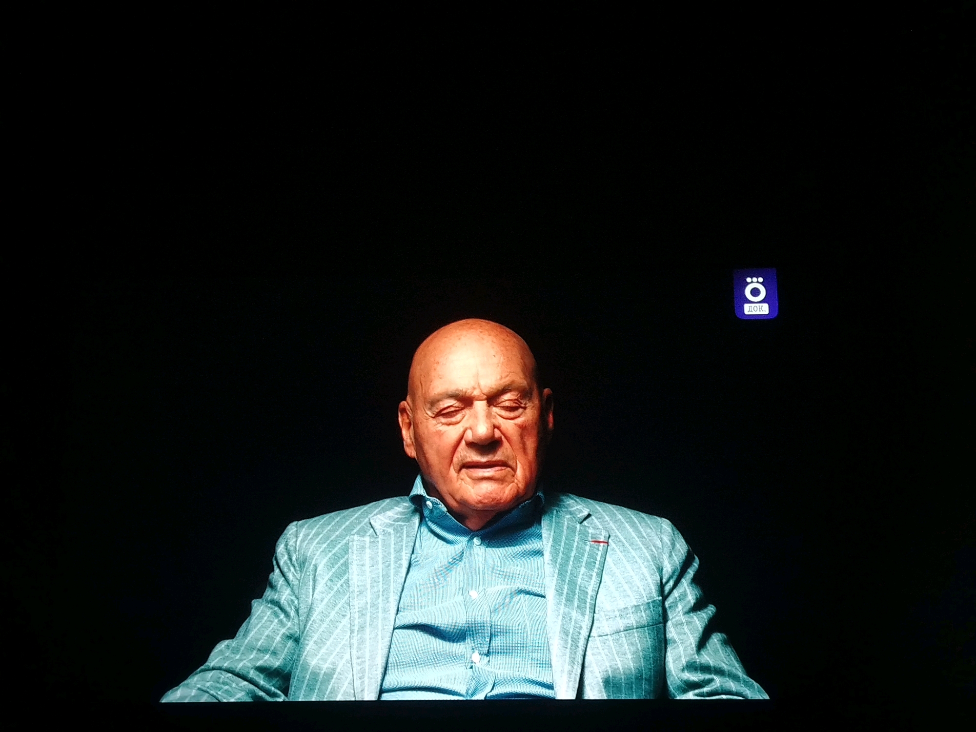 A close-up, head-and-shoulders shot captures an older Caucasian man with his eyes closed, seated against a black background. He has a bald head, wrinkles, and a light complexion. He is wearing a light blue collared shirt with a subtle pattern and a pale mint green pinstripe suit jacket. To the upper right of the frame, a small purple square logo with white text is visible. The lighting is focused on the man, creating a dramatic contrast with the dark surroundings.