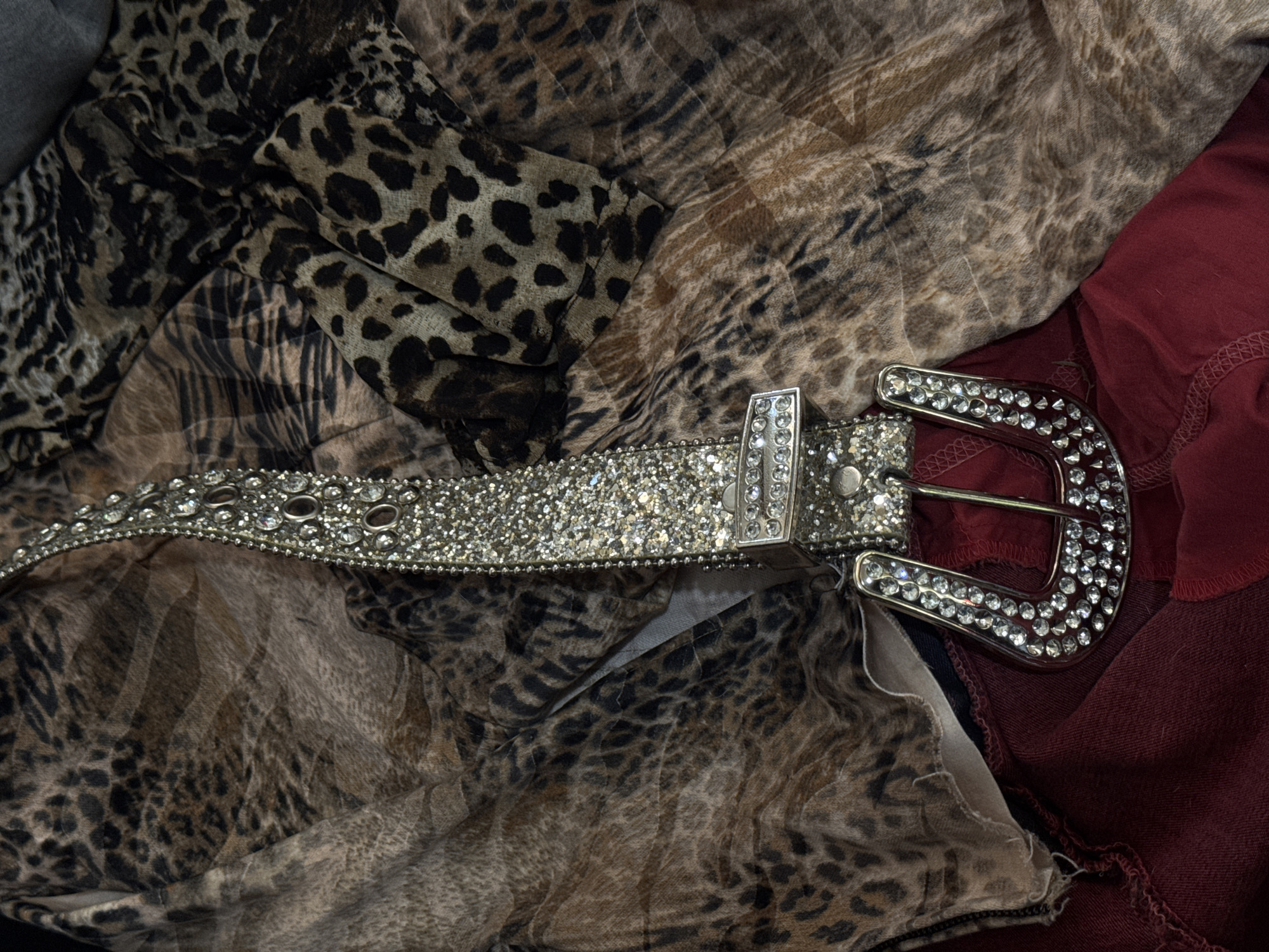 The scene depicts a highly embellished belt featuring numerous clear rhinestones or crystals, laid upon an arrangement of fabrics. The belt, primarily silver-toned, curves across the frame with its large, similarly crystal-adorned buckle positioned in the lower right quadrant. Beneath and around the belt are multiple sections of fabric displaying a brown and black leopard print pattern, appearing folded and layered. A piece of dark red fabric is partially visible along the bottom edge of the frame, underneath the patterned fabrics and the belt. The items are stationary, implying an indoor setting. No individuals, actions, or interactions are present. The location is Sofia, Bulgaria.