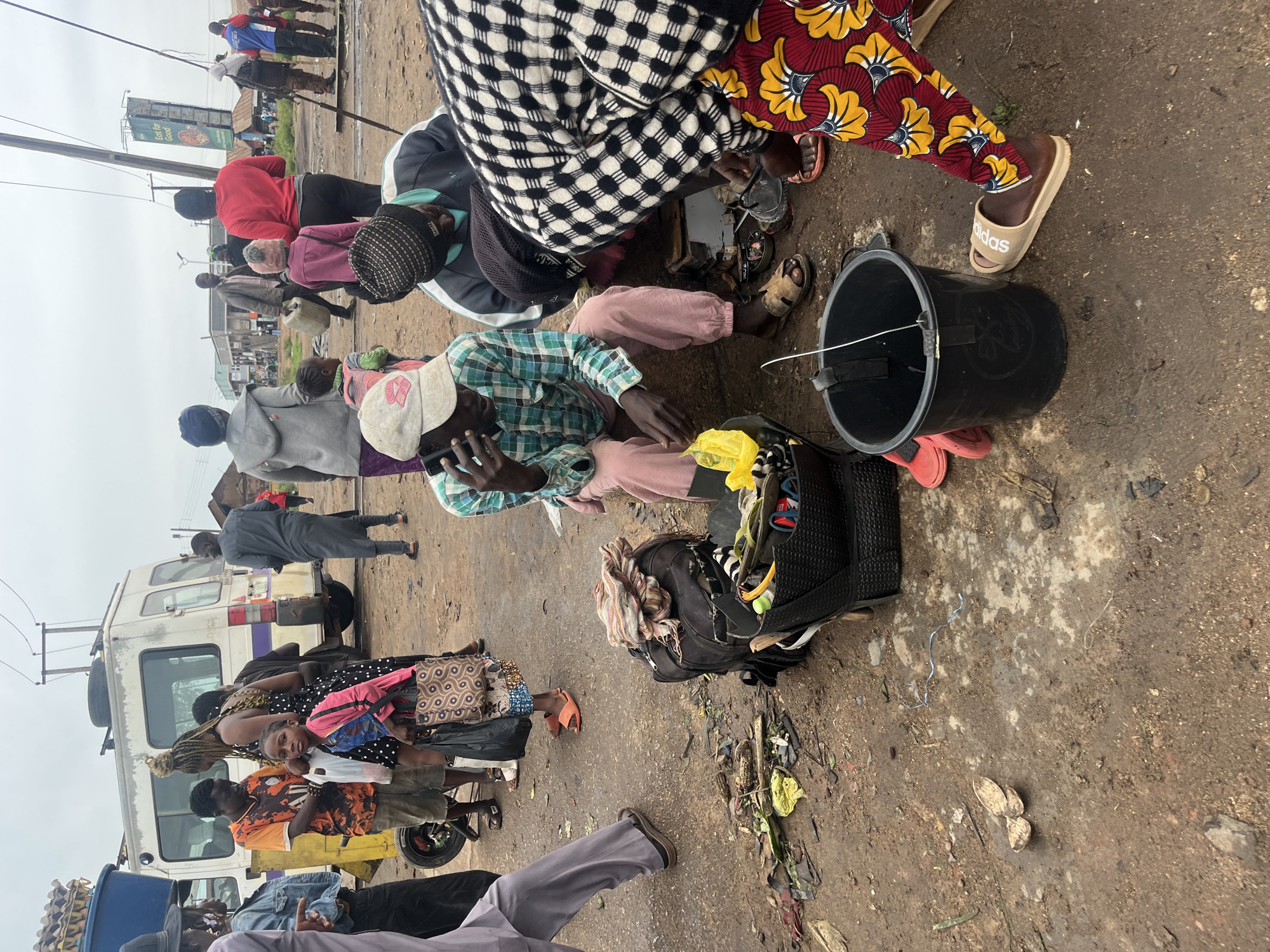 The image depicts an outdoor scene in what appears to be a market or roadside area in Bukuru, Nigeria. The ground is dirt and somewhat wet, with scattered debris. A white bus or van is parked in the background, and a lamppost with overhead wires is visible.
In the foreground, several people are present. A young boy in a pink jacket and black and white polka dot dress is holding a bag, looking towards the camera. To his left, a man in an orange and black patterned shirt is also holding bags. A woman with braided hair is standing next to them.
Further into the scene, a man wearing a green and blue plaid shirt and pink pants is seated, holding a phone to his ear. Beside him, another man in a black and white checkered garment is looking down. A black bucket with a white string hanging from it sits on the ground near the seated man. Another person, wearing a red and yellow patterned fabric on their legs, is also visible.
Various objects are scattered around, including bags, a black backpack, and what appear to be some food items or shells on the ground. The overall impression is of a busy, informal public space.