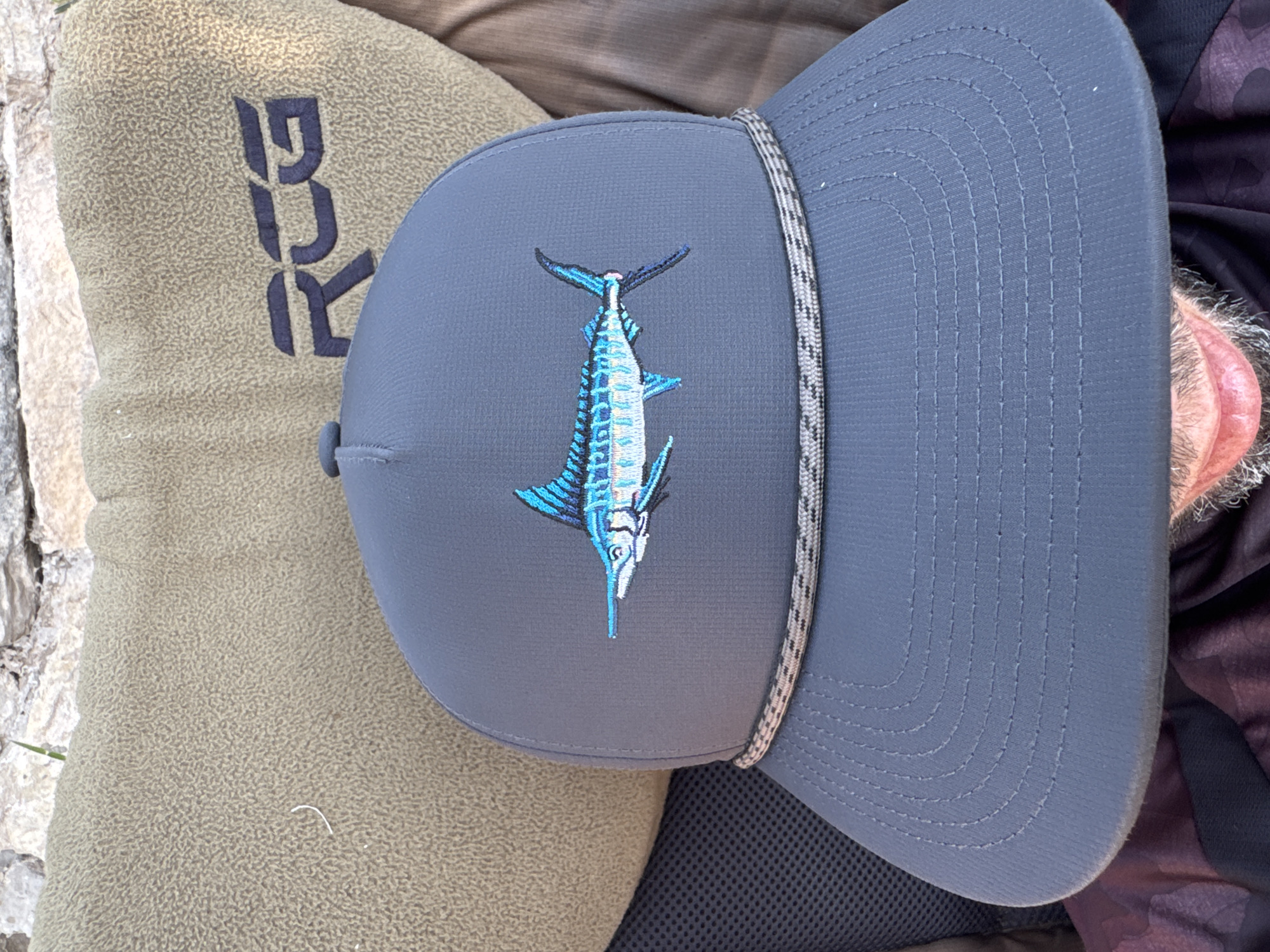 The image shows a close-up, eye-level, outdoor shot of a person wearing a gray baseball cap with a marlin embroidered on the front. The cap has a rope detail across the brim. Behind the cap, a khaki-colored object with a dark blue logo that appears to be "RCG" is visible. The background includes a stone wall and a hint of foliage. The lower part of the image shows the person's mouth and chin with a beard. The lighting suggests it is daytime.
