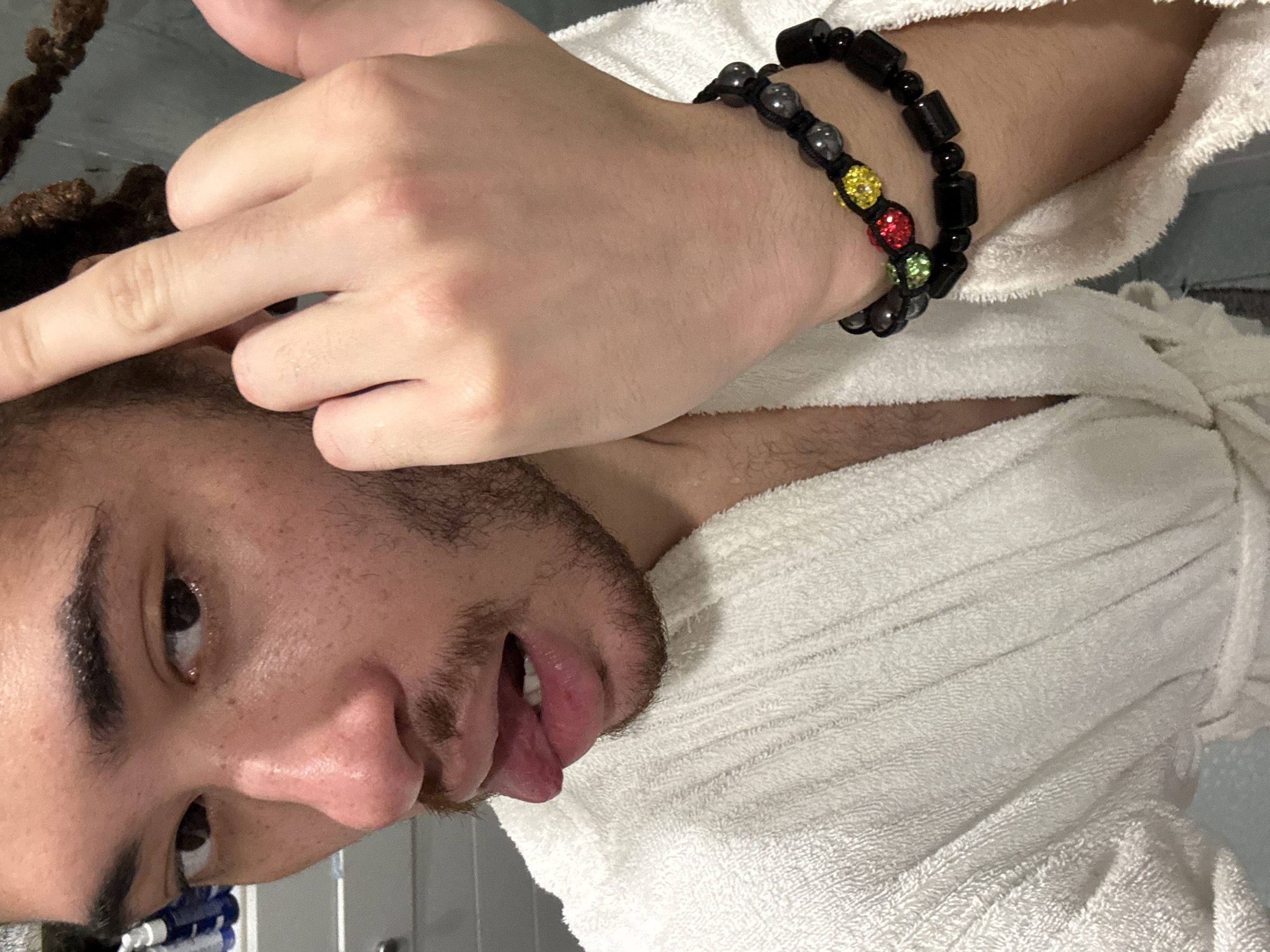 A close-up, low-angle shot depicts a man in a white terrycloth robe. His face is partially visible, showing facial hair, dark eyebrows, and one visible eye. A hand with a dark beaded bracelet rests on his forearm, which is also visible. The man's mouth is slightly open, and his tongue is partially visible. The background is out of focus. There's no information to suggest the location is Les Ulis, France.