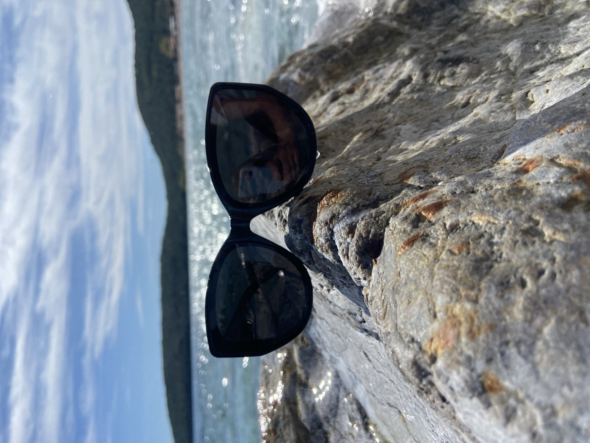 Sunglasses rest on a light gray rock. The rock is textured with variations in tone. The backdrop is a body of water shimmering in the sunlight with trees in the distance. The location is near Prijevor, Montenegro.