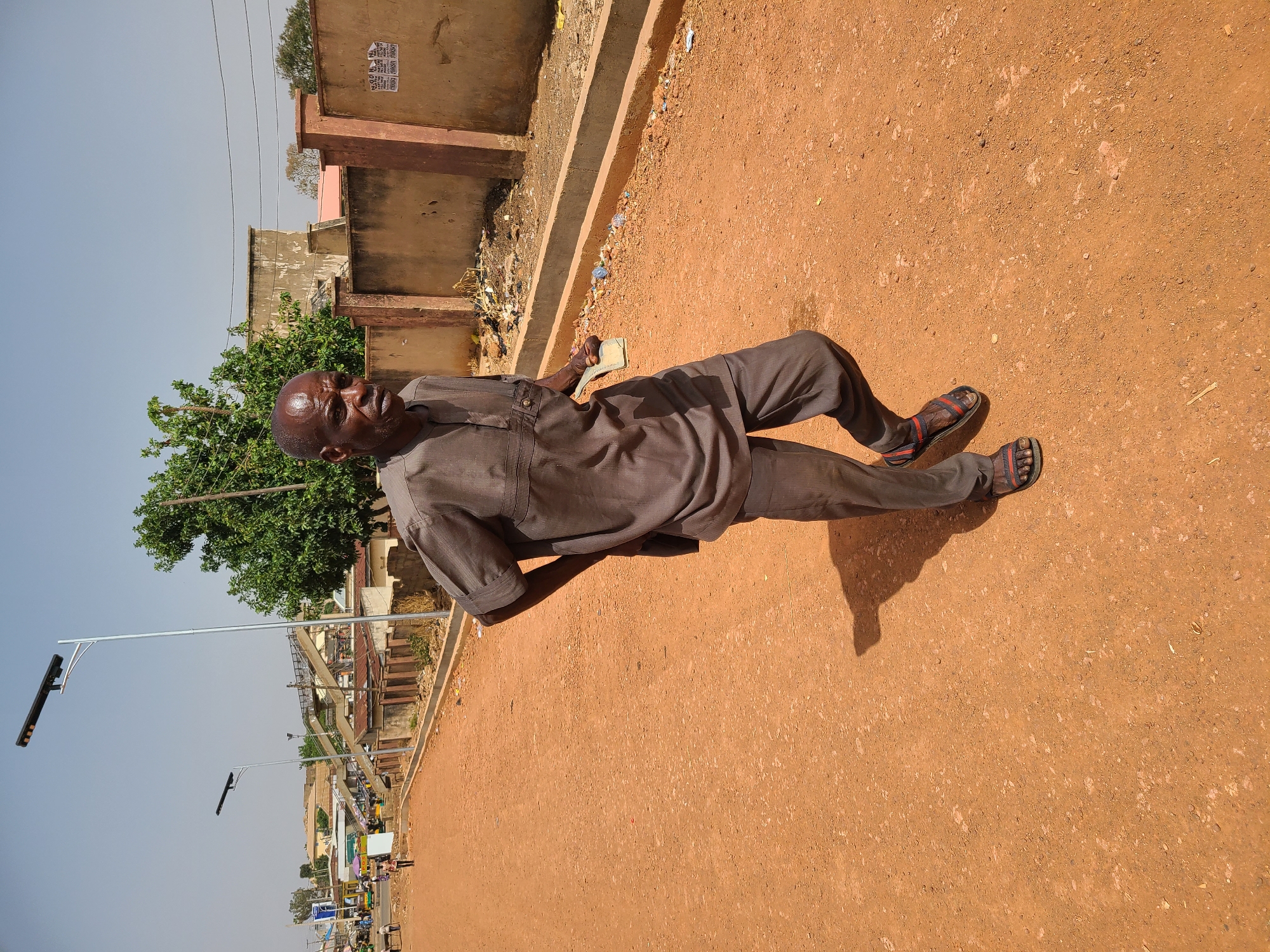 An elderly man lies on his side on a reddish-brown dirt ground in Jos, Nigeria. He wears a grey, short-sleeved button-down shirt and matching trousers, with sandals on his feet. He holds a light-colored rectangular object in his right hand. Behind him is a low, concrete wall with debris, and a green tree is visible nearby. In the distance, streetlights and buildings can be seen on what appears to be a street. The sky is light blue.