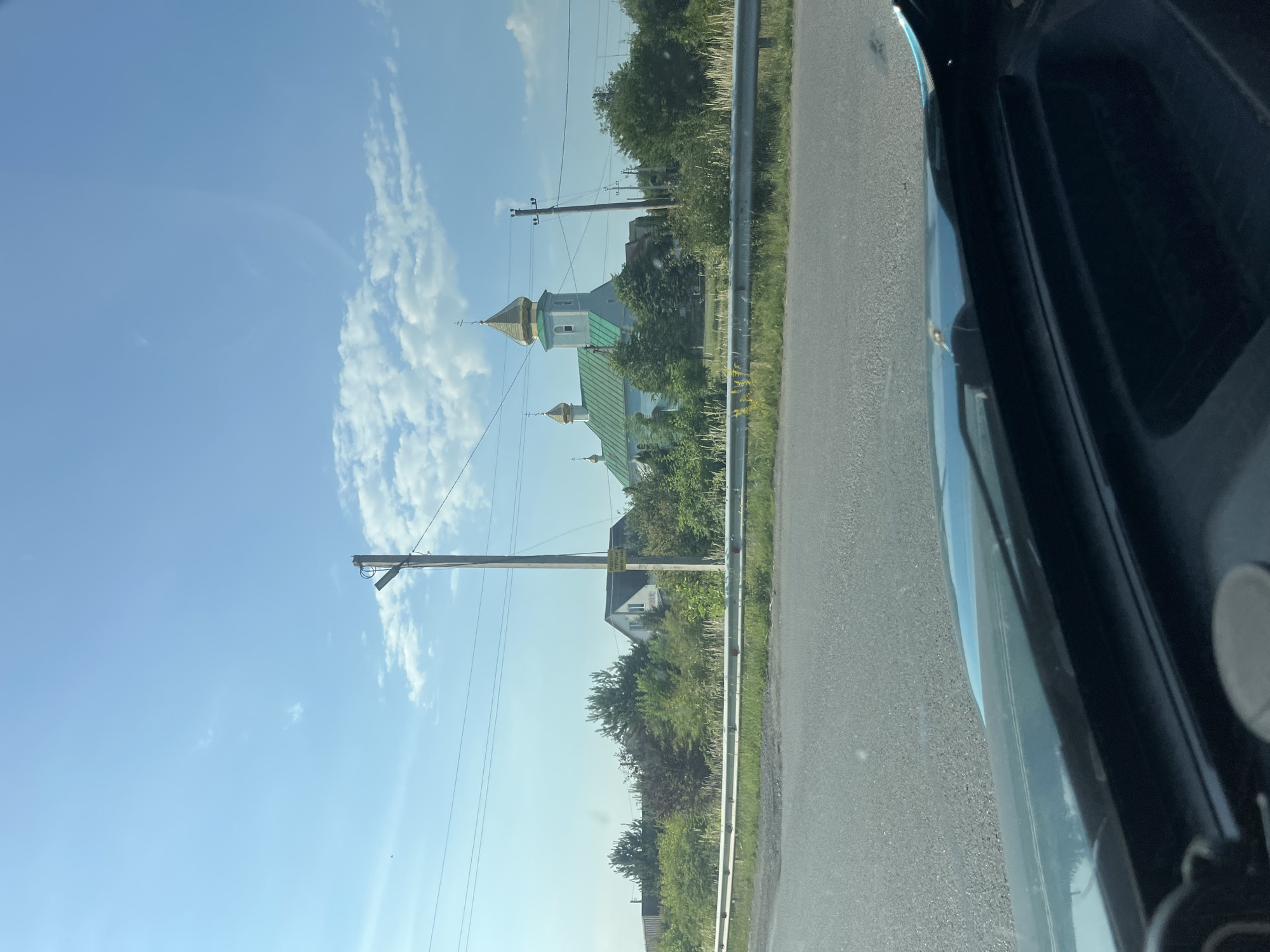 In the city of Baranyntsi, Ukraine, a blue and green church with a golden dome stands amidst lush greenery under a bright blue sky dotted with scattered white clouds. The church, with its distinct architectural style, is flanked by trees and houses, suggesting it is part of a residential area. A metal guardrail runs along the side of a paved road in the foreground, indicating a highway or main thoroughfare. The view appears to be captured from inside a vehicle, as the dashboard and windshield are visible at the bottom of the frame. Utility poles with power lines are scattered across the landscape, connecting to buildings and traversing the open sky. The overall scene depicts a serene, rural setting on a clear day.