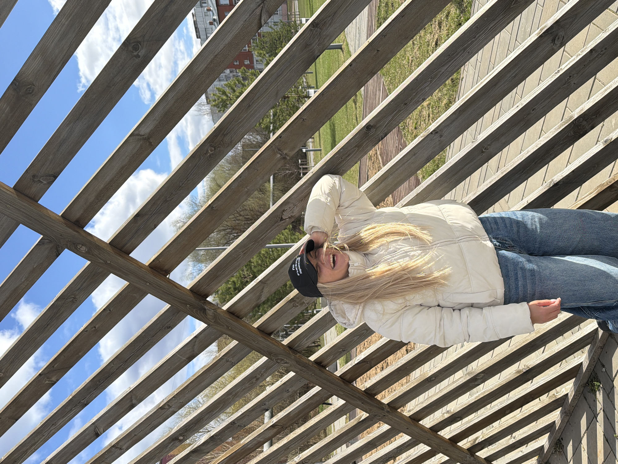 A blonde woman with fair skin is lying on a wooden pergola. She is wearing a black baseball cap, a light-colored puffer jacket, and blue jeans. The cap has white lettering. She is smiling and looking toward the camera. Through the pergola slats, the sky with scattered clouds is visible. In the background, there are trees and buildings, typical of an urban park or recreational area in Votkinsk, Russia. The pergola casts shadows onto the ground.