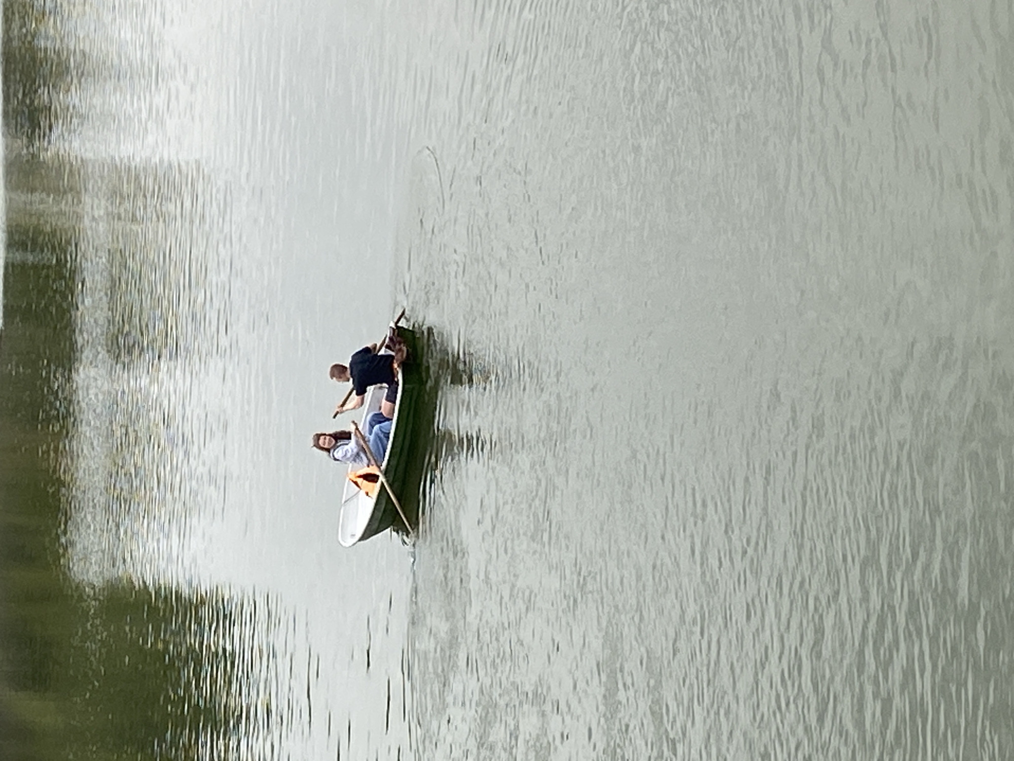 In an outdoor, aerial shot, two people are in a rowboat on a calm body of water. The boat is green and white, with a red life vest visible in the front. The person in the back, a male wearing a dark t-shirt and dark shorts, is rowing. The person in the front, a female wearing a light-colored hooded sweatshirt and blue jeans, is looking towards the viewer and smiling. The water reflects the trees and sky, creating a dappled effect. The overall scene depicts a leisurely day on the water.