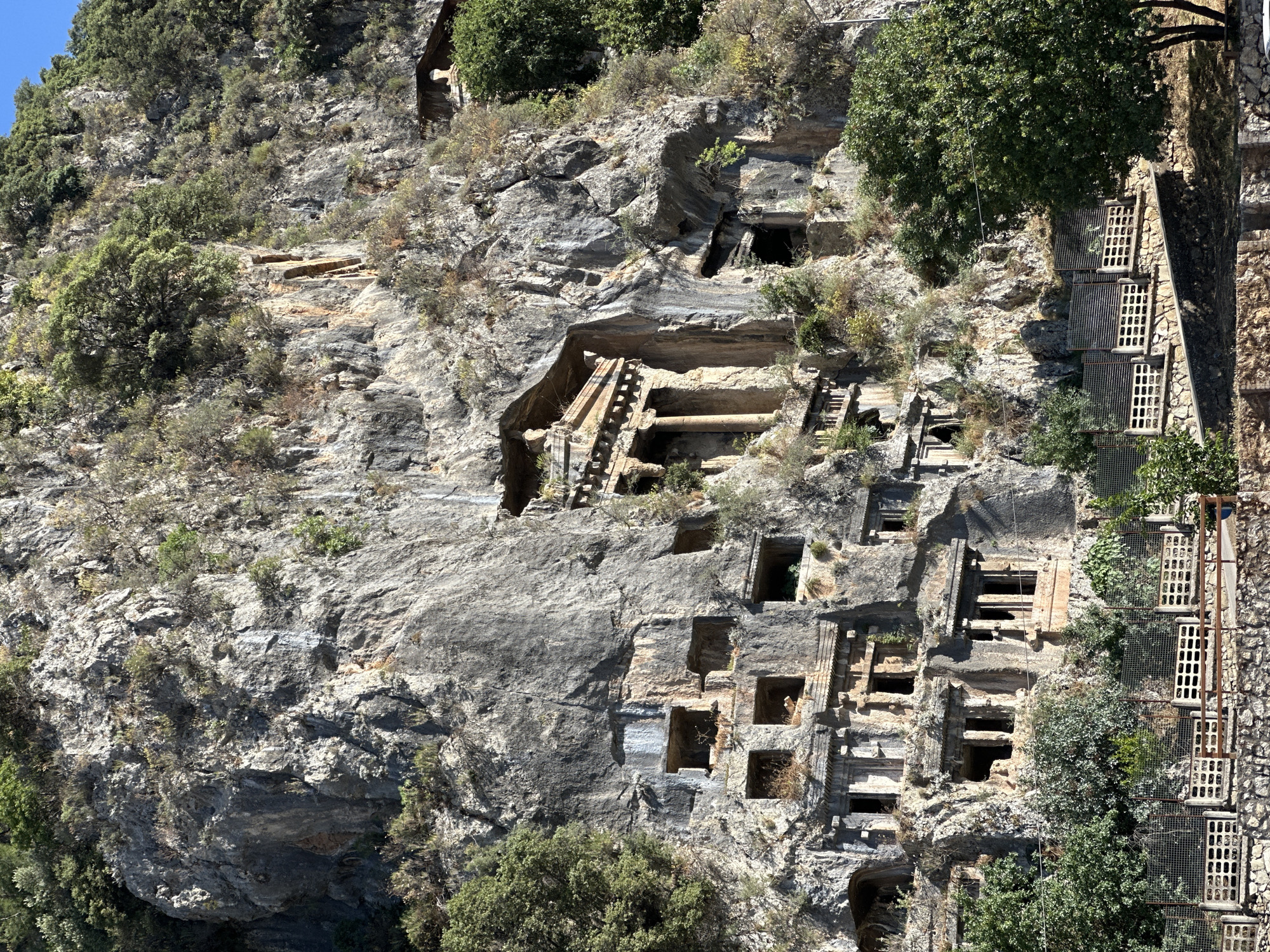 A steep, rugged cliff face, predominantly gray and rocky, is densely carved with numerous ancient rock-cut tombs. These tombs vary in design; some feature ornate, multi-story facades with steps and platforms, while others appear as simpler square or rectangular openings. Many of the tomb entrances are dark, suggesting deep chambers within the rock. Patches of green vegetation, including small trees and bushes, cling to the cliff face and grow between the tomb structures, highlighting the passage of time and integration with nature.
On the far right, a more modern structure is visible, consisting of a stone wall and a series of vertically stacked, gridded metal platforms or enclosures, possibly indicating a modern viewing platform, walkway, or safety netting for visitors. Sunlight illuminates the entire scene, casting distinct shadows and suggesting a bright, clear day, likely midday or early afternoon. The site is an outdoor archaeological and historical location, characteristic of the Lycian rock tombs found in Fethiye, Turkey. No specific activity is taking place, but the scene speaks to the ancient human endeavor of carving elaborate burial sites into natural landscapes. There is no visible text.
