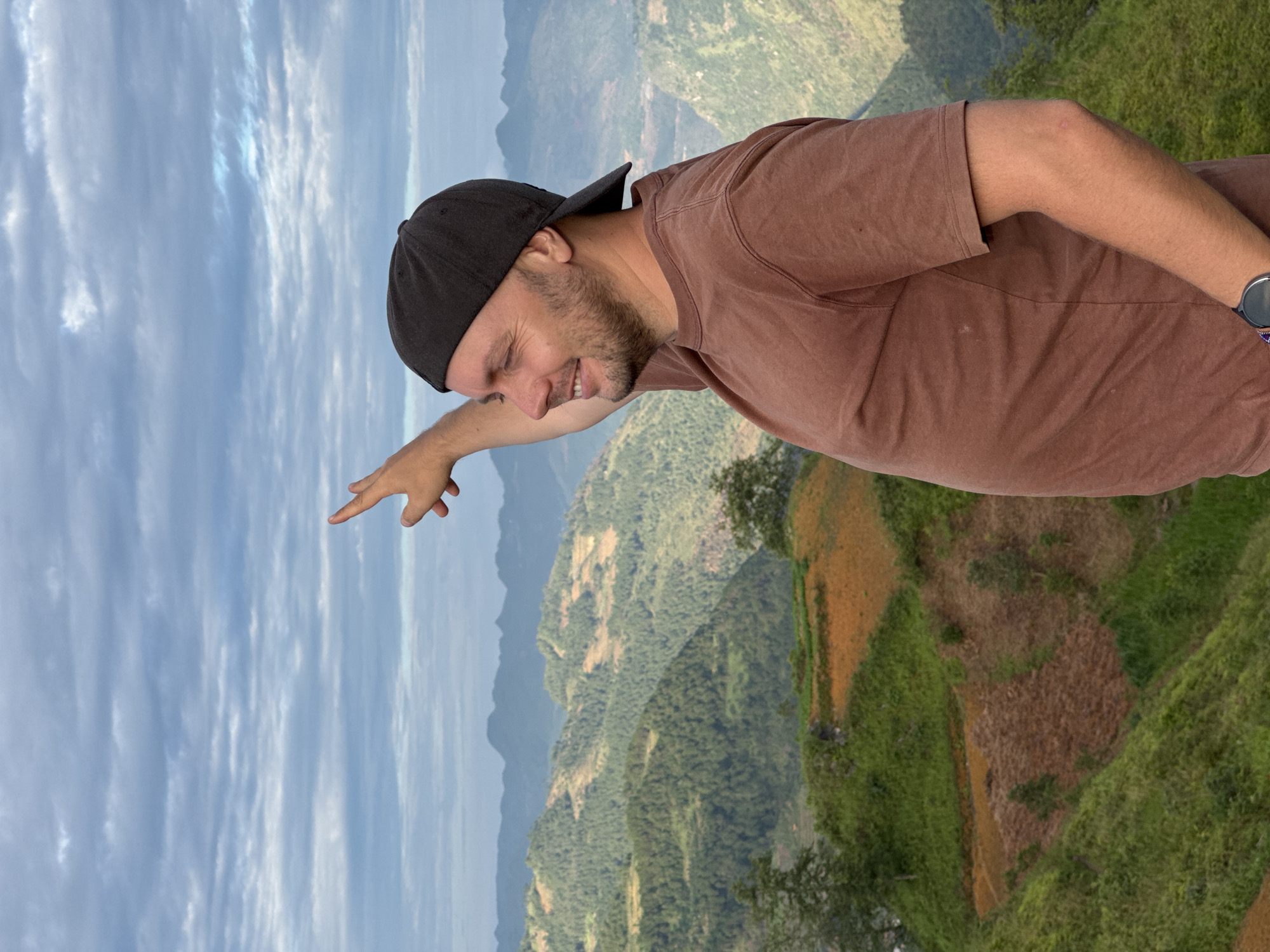 A man wearing a black baseball cap and a brown t-shirt is standing on a mountain ridge in Lũng Cú Commune, Vietnam. He is pointing with his left hand towards the sky, which is partly cloudy. The background features a mountainous landscape with green vegetation and terraced fields.