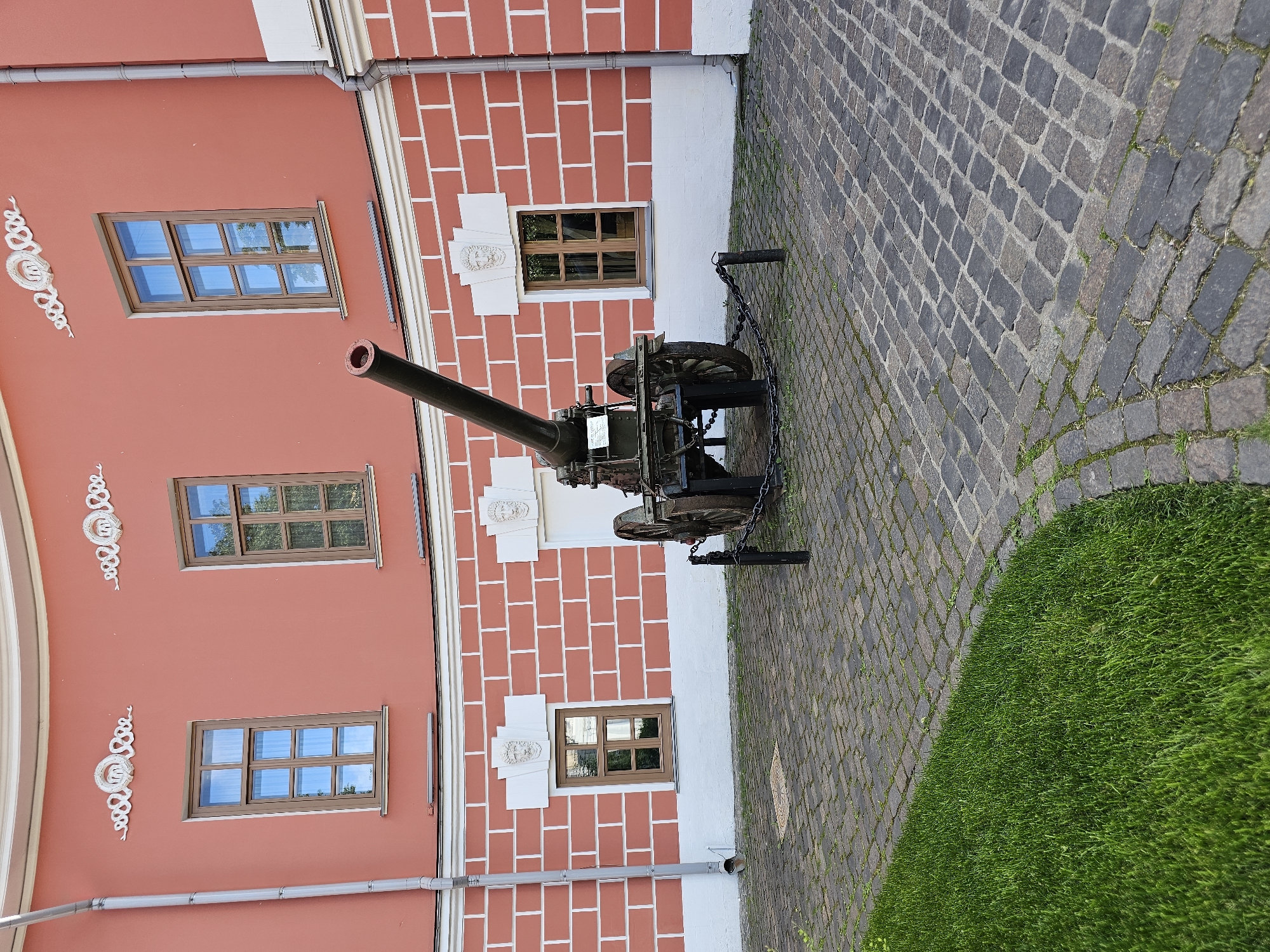 The image shows an old cannon positioned in front of a red brick building with white trim. The cannon is on a wheeled carriage and is surrounded by a low chain fence. The building has several windows and decorative elements. The ground is paved with cobblestones, and there is a patch of green grass on the left side of the image. This scene is located at the National Military History Museum of the Armed Forces in Moscow, Russia.