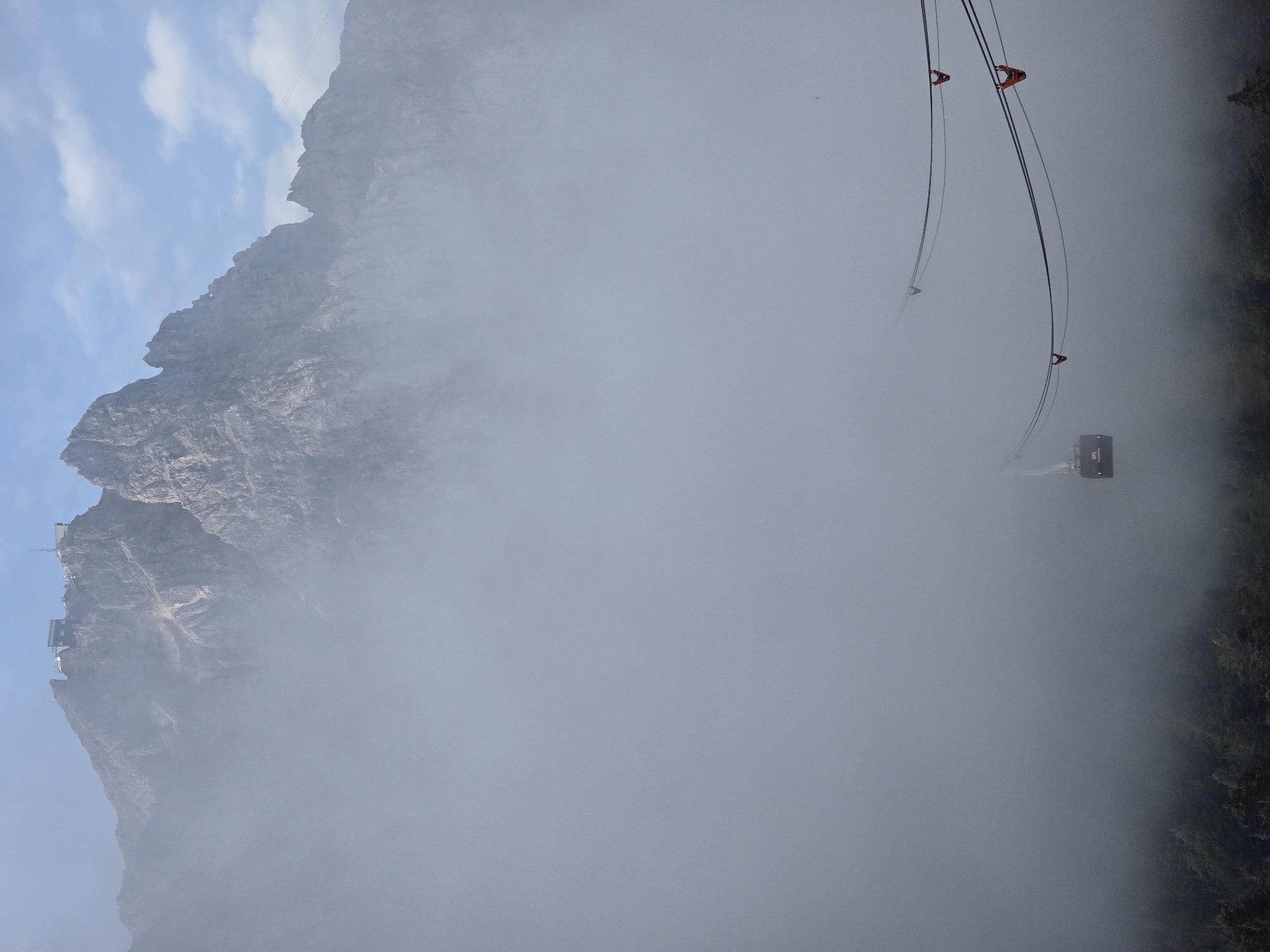 The image depicts the Zugspitze mountain in Grainau, Germany, partially obscured by thick fog. At the summit, a building complex, likely a hotel or observation deck, is visible against a clear blue sky with scattered clouds. In the mid-ground, a cable car, also known as a gondola, traverses the misty atmosphere, suspended by cables. The lower portion of the scene shows a dense forest, its trees silhouetted against the fog. The cable car appears to be in motion, suggesting transportation up or down the mountain. No people are discernible in the image. The overall atmosphere is one of mystique and grandeur, with the fog adding a dramatic element to the mountainous landscape.