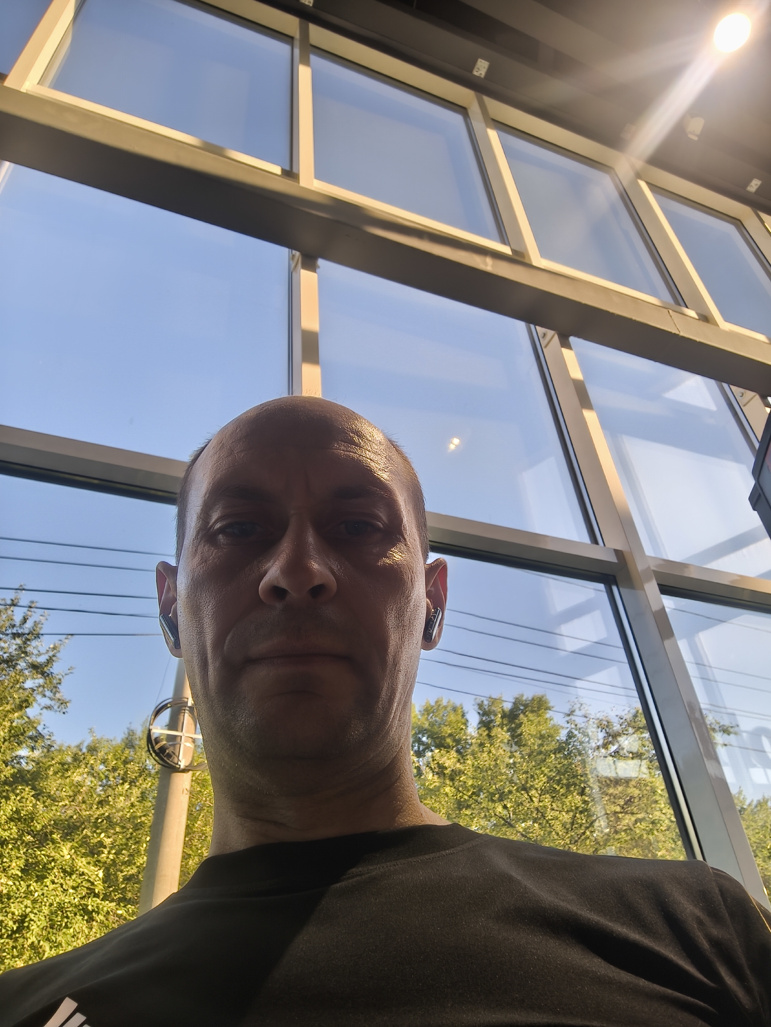 This is a low-angle, close-up, self-taken photograph of a man's face and upper torso, taken outdoors. The man is bald and wearing a black t-shirt and wireless earbuds. He is looking directly at the camera with a neutral expression.
Behind him is a large window with a light-colored frame, divided into multiple panes. The sky is visible through the windows, appearing bright blue with a few faint reflections. Sunlight is streaming through the upper part of the window, creating a bright glare.
Outside the window, on the left side of the frame, are trees with green foliage. In the lower left corner, part of a utility pole with a circular fixture is visible. The overall lighting suggests it is daytime.
The location of this photograph is not specified in the image. Therefore, it is not possible to determine if it was taken in Komsomolsk-on-Amur, Russia.