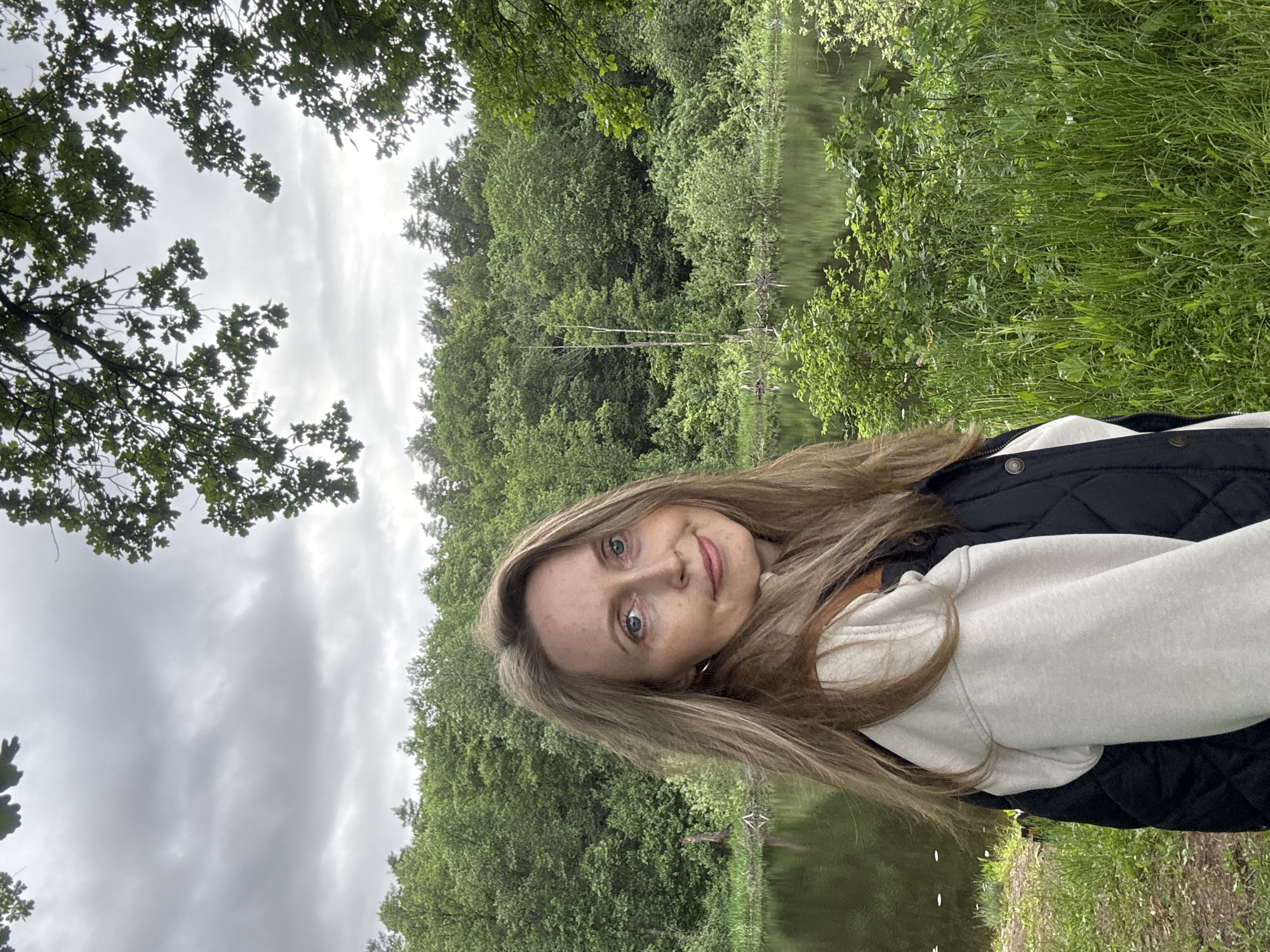 The image displays a woman outdoors, positioned in the foreground against a natural backdrop. She has long, light brown hair, blue eyes, and freckles, looking directly at the camera with a subtle smile. She is wearing a light-colored hooded sweatshirt beneath a dark, quilted jacket. A gold hoop earring is visible in her right ear. Behind her, a calm body of dark water, likely a pond or small lake, reflects the overcast sky and surrounding vegetation. The background features a dense forest composed of various deciduous and coniferous trees with lush green foliage. Several thin, light-barked trees, some appearing bare, stand at the water's edge on the far bank. Overcast grey clouds dominate the sky. In the upper left and right corners, leafy tree branches extend into the frame. The immediate foreground shows green grass and low-lying vegetation. There are no discernible landmarks or identifying features within the image to confirm the specific location as Chaikovsky, Russia.