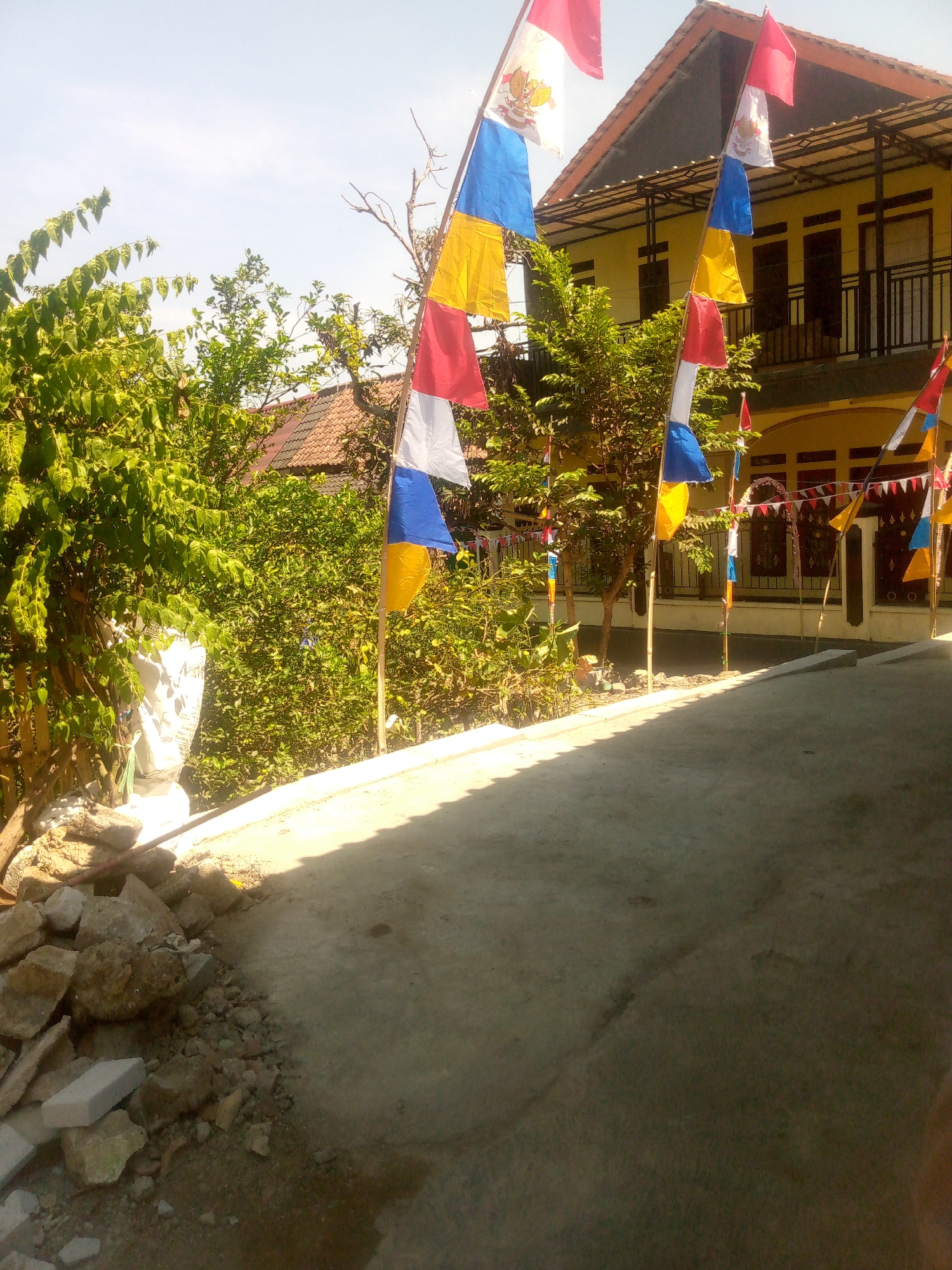 In the city of Giriasih, Indonesia, on a clear day, a row of flags flutters in the breeze. The flags are small and triangular, with alternating colors of red, white, blue, and yellow. They are strung together and attached to bamboo poles, lining a paved walkway in front of a yellow building. The building has multiple windows and a balcony. To the left of the building, there is dense green foliage and trees. In the foreground, a pile of rocks and debris sits on the edge of the walkway. The scene appears to be decorated for a festive occasion.