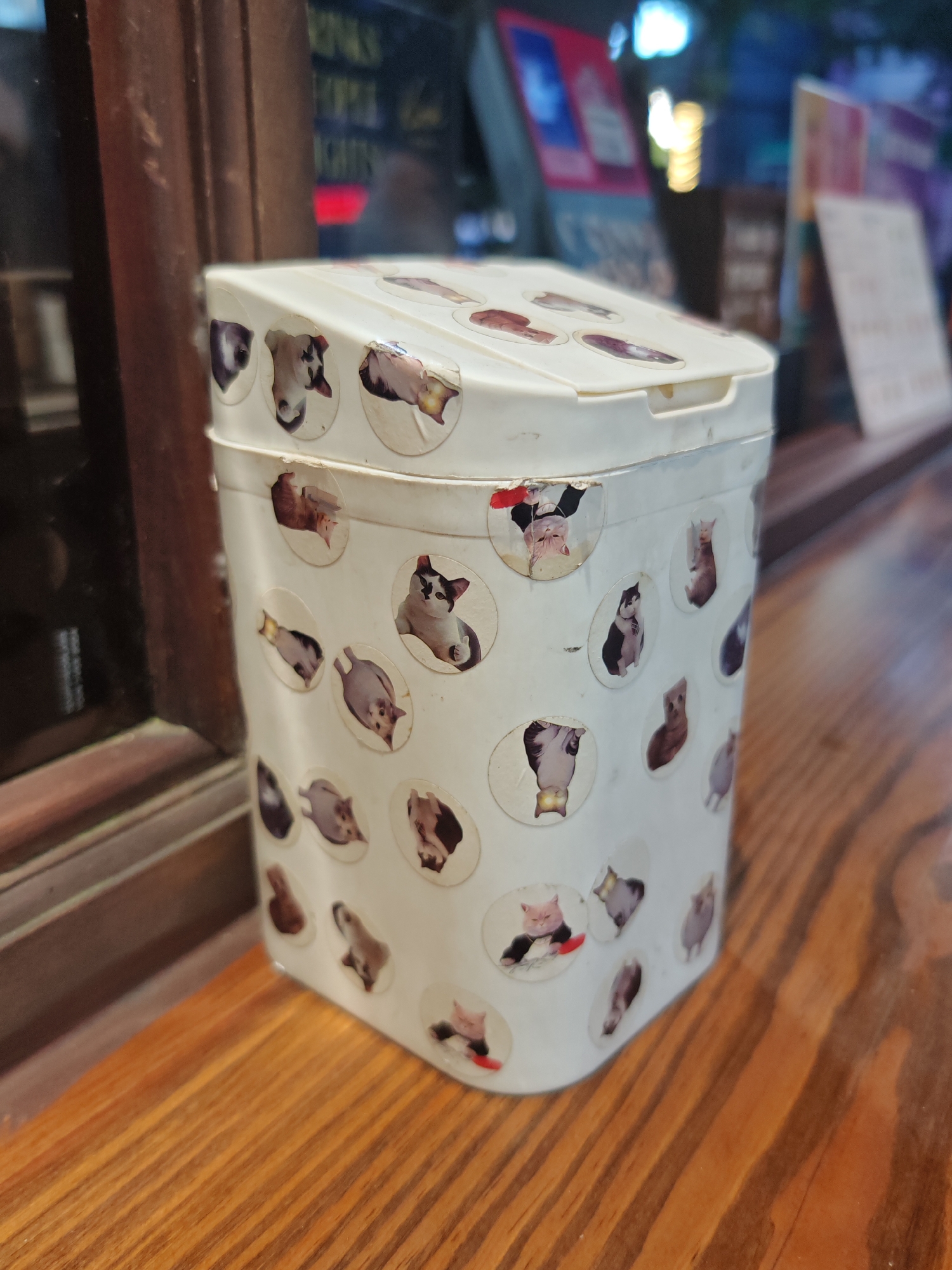 A close-up, low-angle shot captures a white, cylindrical container with a hinged lid, adorned with numerous circular stickers featuring cartoonish depictions of cats and cows. The container is positioned on a wooden surface, likely a table or shelf, with a dark wooden frame visible to the left. In the blurred background, shelves are lined with books and other items, illuminated by artificial lights, suggesting an indoor setting such as a shop or library. The overall scene is static, with no discernible action or interaction. The image does not provide any specific information to determine its location within Nha Trang Ward, Vietnam.