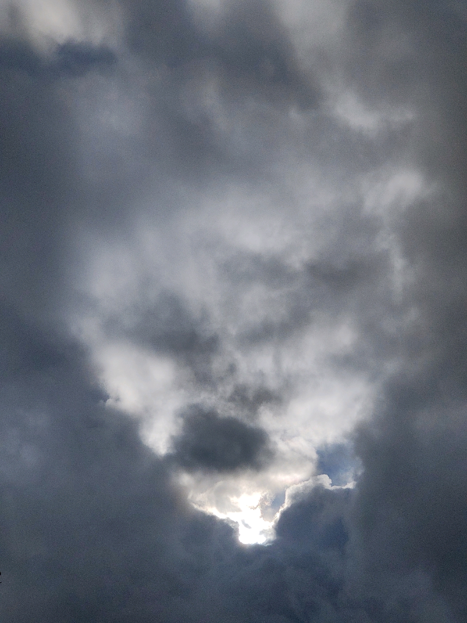A dramatic, overcast sky over Warmond, Netherlands. The clouds are dark and heavy, suggesting an impending storm. A bright break in the clouds reveals a patch of blue sky and the sun, casting a powerful light that pierces through the gloom. The light creates a halo effect around the edges of the clouds, highlighting their texture and depth. The overall impression is one of a powerful and dynamic natural event, with the light representing hope amidst the darkness.