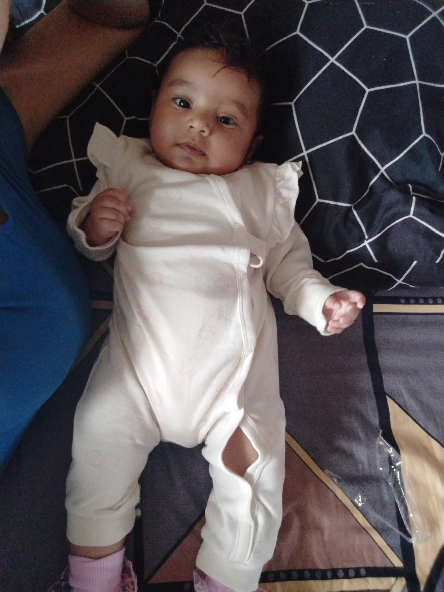 A baby lies on a patterned blanket. The baby is wearing a white romper with pink patterns and ruffled shoulders. The romper has an opening at the leg. The baby is lying on its back with both arms extended and looking directly at the camera. The blanket beneath the baby has a black and white geometric pattern in the top left corner, transitioning into a brown and grey striped pattern with gold accents towards the bottom right corner. To the left of the baby is a person wearing a blue shirt. The location and city are unknown.