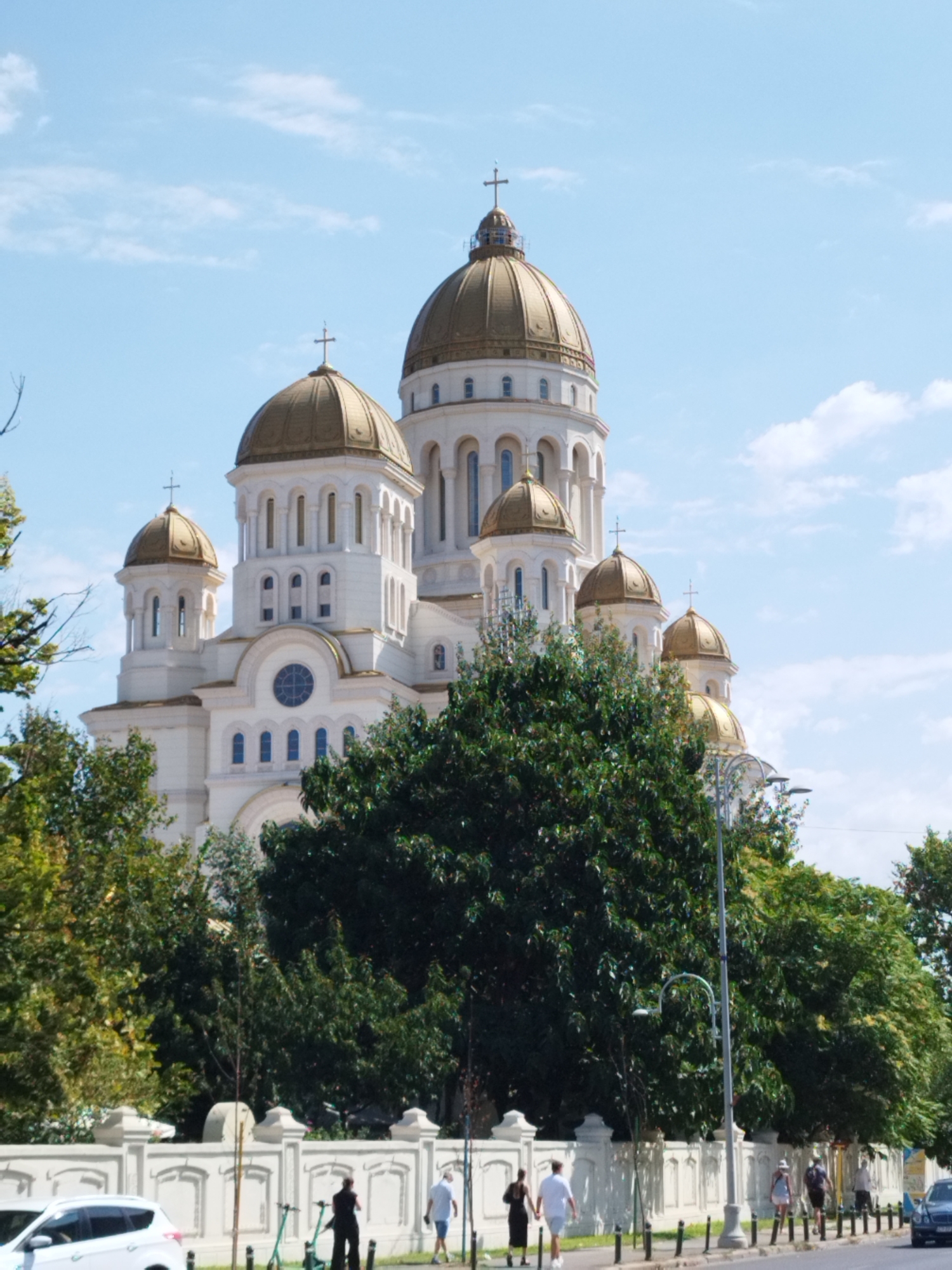 The image captures a view of the Saint Nicholas Cathedral in Bucharest, Romania, on a partly cloudy day. The grand cathedral, with its white facade and multiple golden domes topped with crosses, dominates the upper half of the frame. Lush green trees partially obscure the lower portion of the building. In the foreground, a white decorative wall lines the street, behind which a white car is parked on the left. A streetlamp stands prominently in the middle of the sidewalk. Several individuals are visible walking along the sidewalk: a woman in a black dress and a man in a white shirt and shorts are walking together on the left, while a man in a white shirt and shorts and a woman in a white top and shorts are walking together further down the street to the right. Two green electric scooters are parked near the wall. A black car is parked on the far right side of the image, and other cars are visible in the distance. The sky is a bright blue with scattered white clouds.