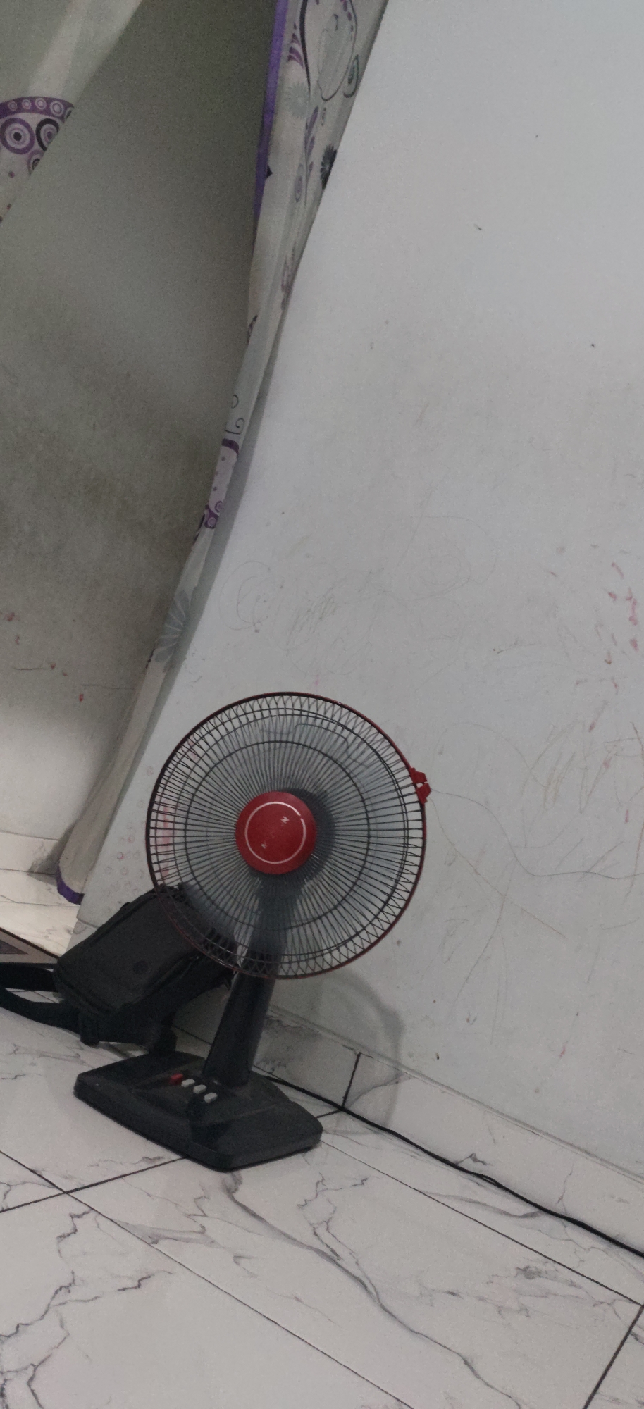 A black standing fan with a red central dial is positioned on a marble-patterned floor in front of a white wall. The wall has faint red and gray markings, possibly from children's drawings or scuffs. To the left of the fan, a black bag rests against a fabric curtain with a circular purple and gray pattern. The scene appears to be indoors, likely in a residential setting, in West Jakarta, Indonesia. The lighting is dim, casting a shadow from the fan onto the wall. There are no people visible in the image.
