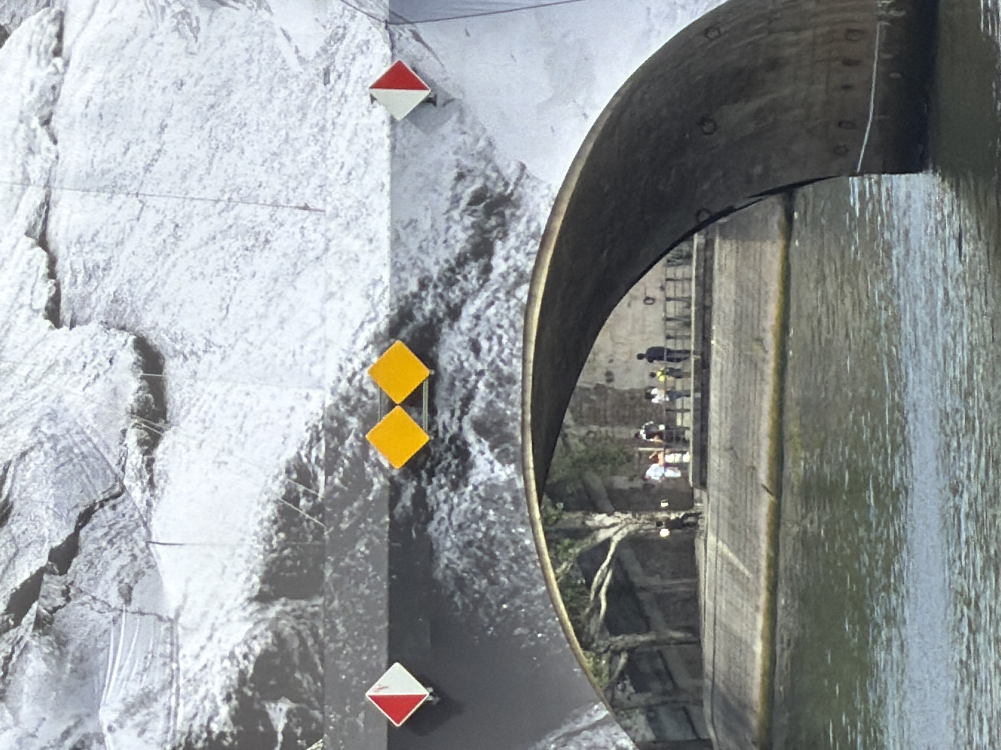 The image captures a view of the Seine River in Paris, France, with a bridge arching over the water. On the bridge, there are several diamond-shaped signs. On the left side of the bridge, a red and white diamond-shaped sign is visible. To its right, two yellow diamond-shaped signs are placed next to each other. Further to the right, another red and white diamond-shaped sign is present. Beneath the bridge, a stone embankment runs along the river, and several people are seen standing on the embankment. The water of the Seine appears somewhat choppy. The background of the bridge is a large-scale photograph or mural depicting a rocky, mountainous landscape.