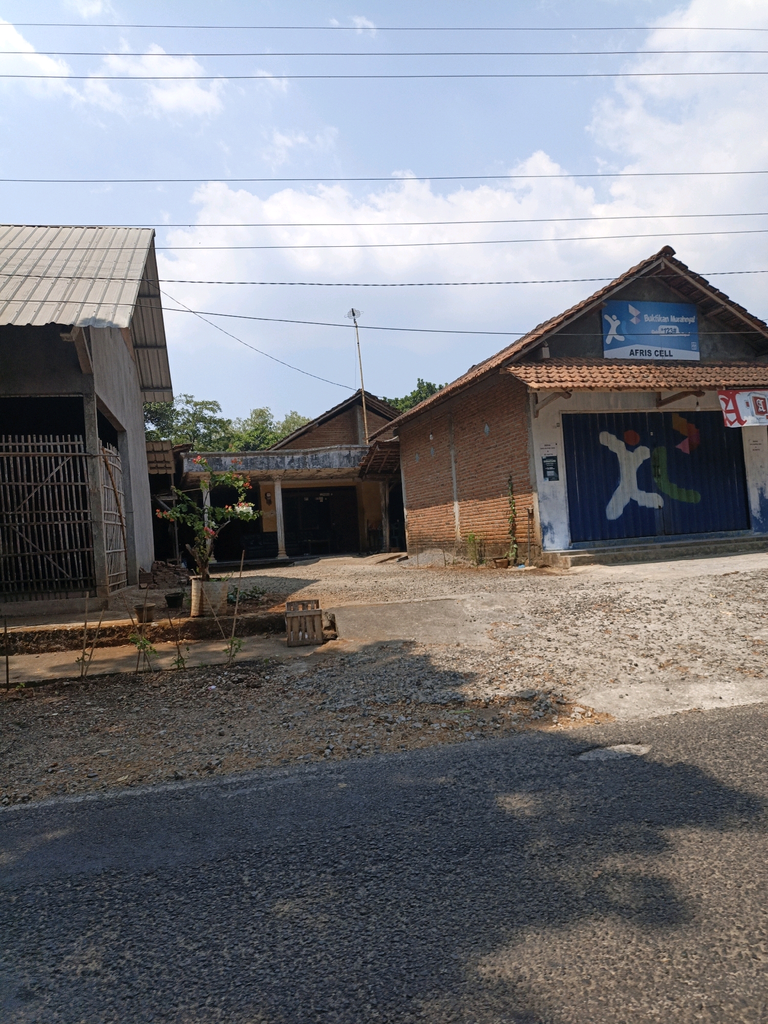The image depicts a street scene in the city of Cikal, Indonesia. The foreground is dominated by an asphalt road with gravel and dirt on the shoulder. In the middle ground, there are several buildings. To the left, a building with a corrugated metal roof and a fence made of bamboo poles is visible. Next to it, there is a smaller building with a tiled roof and an open doorway. To the right, a brick building with a tiled roof features a blue rolling door painted with colorful abstract figures and a sign that reads "AFRIS CELL." Above the door, a banner displays text in Indonesian. In the background, trees and a clear blue sky with clouds are present. The scene is bathed in sunlight, suggesting it is daytime.