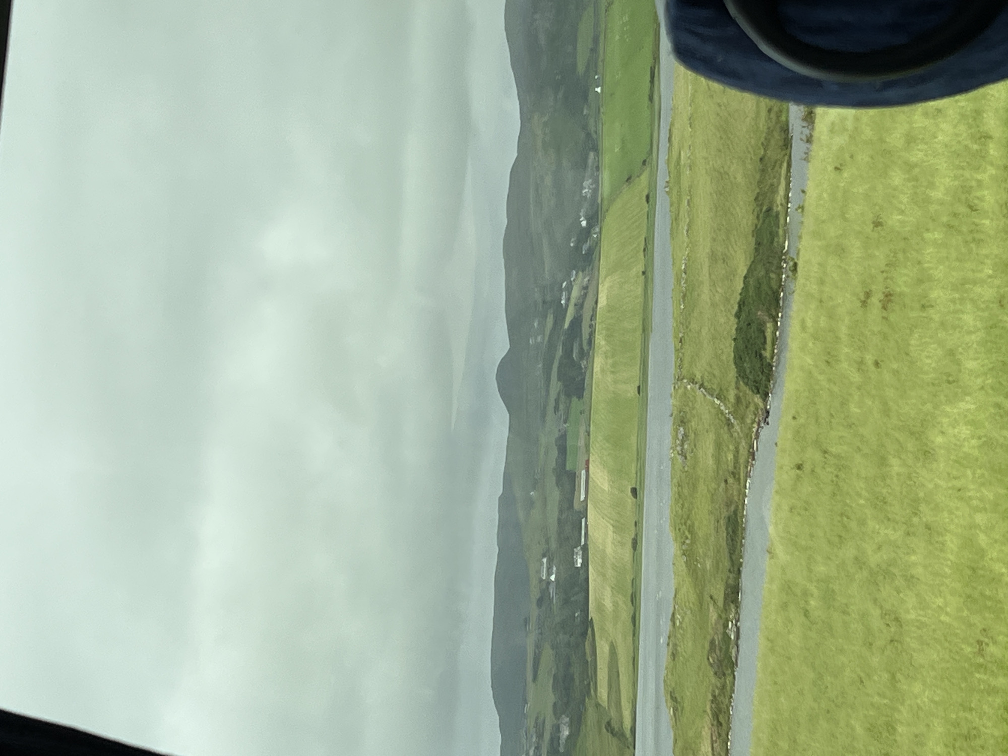 The image displays a landscape view of a rural area, likely in the vicinity of Dunfanaghy, Ireland, under a cloudy sky. In the foreground, a vibrant green grassy field stretches out. A body of water, possibly a lake or inlet, runs through the mid-ground, with grassy banks and some stone features along its edge. Beyond the water, there are more green fields, some appearing cultivated with distinct patterns. Scattered white buildings, indicative of a village or scattered dwellings, are visible on the slopes of rolling hills in the background. These hills are covered in a mix of green vegetation and darker, more rugged terrain, culminating in a hazy mountain range under the overcast sky. A dark blue object, likely a seat or part of a vehicle, occupies the lower right corner of the frame, suggesting the photograph was taken from within a moving vehicle.