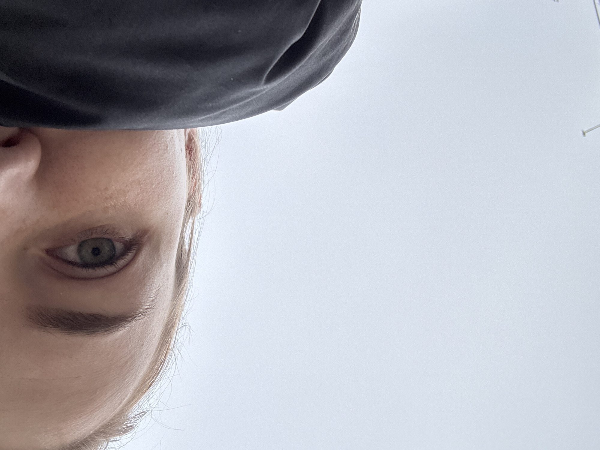 The image shows a close-up view of a person's face from a low angle, with a portion of their black clothing visible at the top of the frame. Only the left side of the face is clearly visible, including the eye, eyebrow, nose, and part of the cheek. The eye appears to be green. The background is a light, overcast sky. There is a small, barely visible structure on the horizon. The location of Krasnodar, Russia cannot be determined from the image.