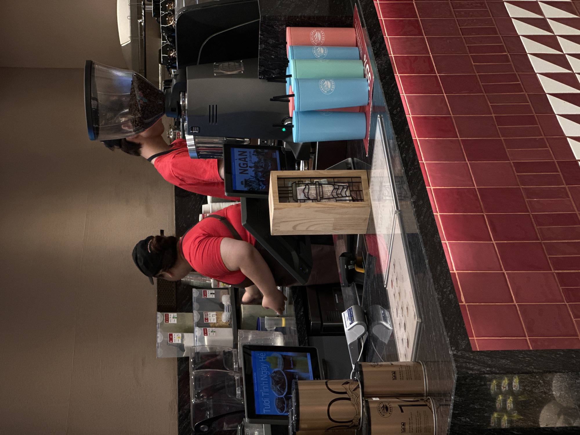 The image depicts an indoor scene within a coffee shop, likely located in the city of Da Nang, Vietnam, given the context of "Highlands Coffee" branding visible on several items.
In the foreground, a black granite countertop is visible, with a portion of the counter's front facade tiled in red. To the left, there are two cylindrical gold canisters with wave-like patterns and text. One canister appears to have "1000" and "CA PHE AU LAC" printed on it. A point-of-sale device is situated on the countertop between the canisters and a touchscreen monitor. The monitor displays a colorful advertisement for iced coffee drinks, with Vietnamese text that includes "Tươi Tình Ngày Hè".
Further back, on the countertop, a row of colorful reusable cups with straws is arranged. These cups are light blue, mint green, and coral pink, and some have a "Highlands Coffee" logo. To the left of the cups, a wooden dispenser with napkins is present.
Behind the counter, a coffee grinder with a clear hopper filled with coffee beans is prominently displayed. Two individuals, presumed to be staff members, are working behind the counter. One, wearing a red shirt and apron, is a woman with dark hair pulled back and is partially obscured as she interacts with equipment. The other, also in a red shirt, is a younger individual whose face is turned away from the camera as they attend to the coffee grinder. In the background, translucent storage bins containing various ingredients, such as white powder and green matcha powder, are visible on shelves. The overall environment suggests a functional coffee shop service area.