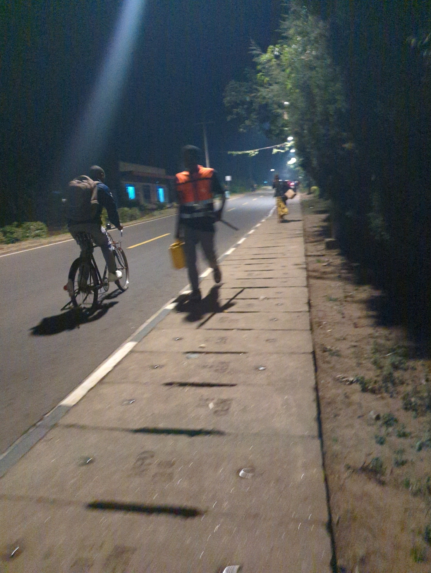 The image shows a nighttime scene on a street in Musanze District, Rwanda. A person is riding a bicycle on the left side of the road, wearing a backpack. Further down the sidewalk, another person walks in an orange safety vest, carrying a yellow bucket. The street is illuminated by streetlights, and trees line the right side of the sidewalk. Buildings are visible in the background. The lighting is dim, creating shadows and a somewhat grainy quality to the image.